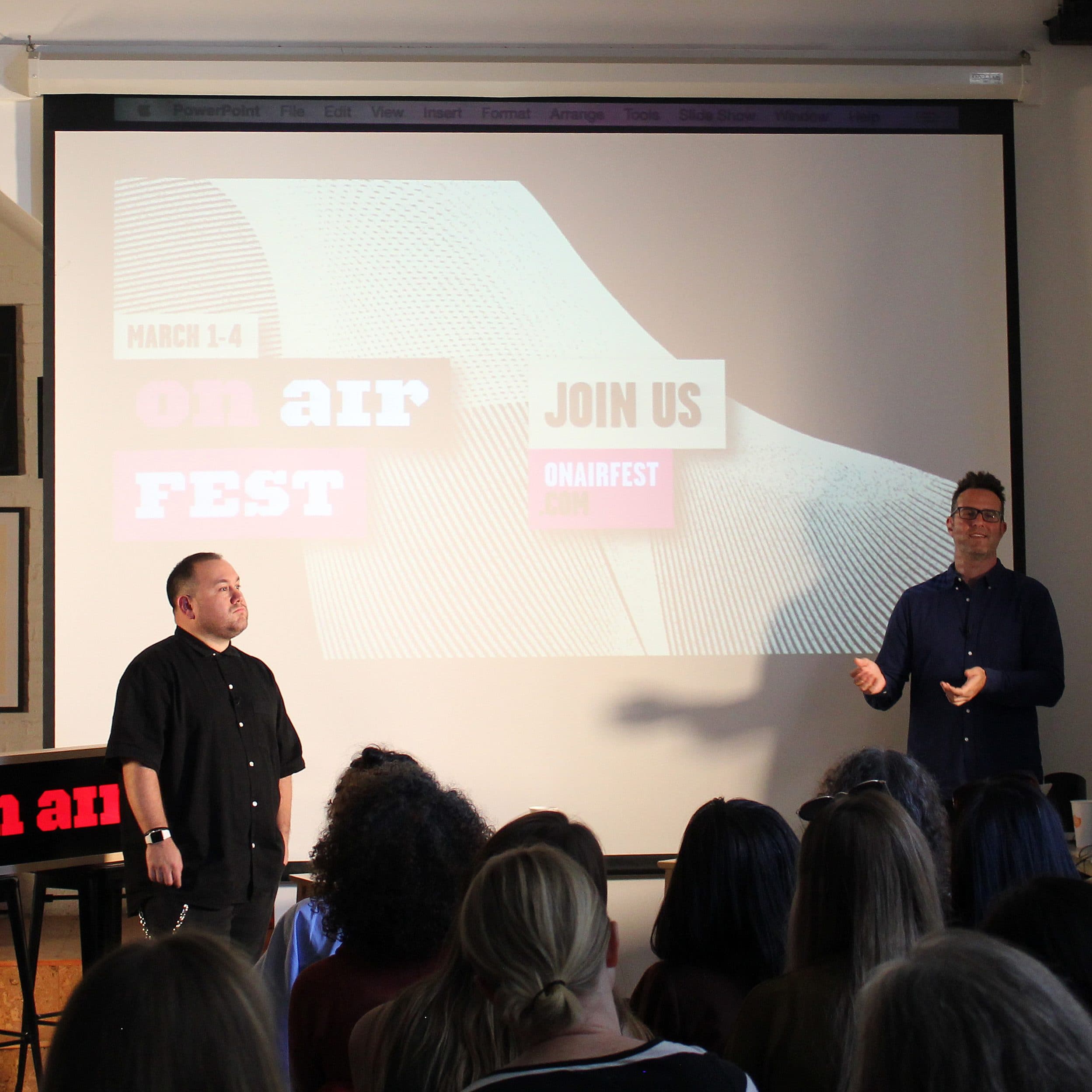 Two men stand in front of an audience in a well-lit room with a large projector screen. The screen displays "March 1-4 On Air Fest" and "Join Us ONAIRFEST" in bold text. A small table with a red "on air" sign is in the foreground on the left.