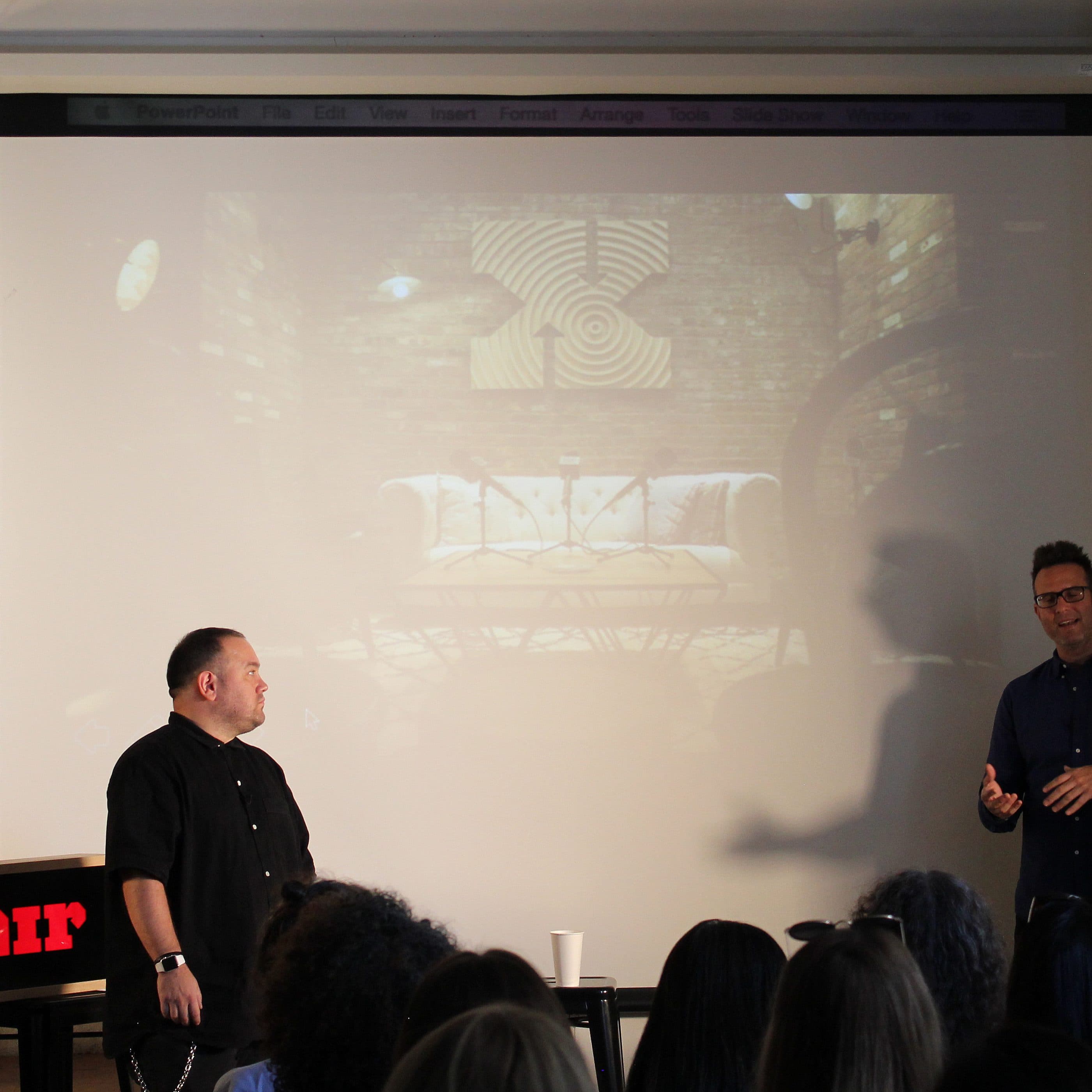 Two men are standing in front of a projection screen giving a presentation. One man on the left is wearing a black shirt and the other on the right is wearing a blue shirt. The room is dimly lit, and a red "on air" sign is visible on a table in front of the audience.