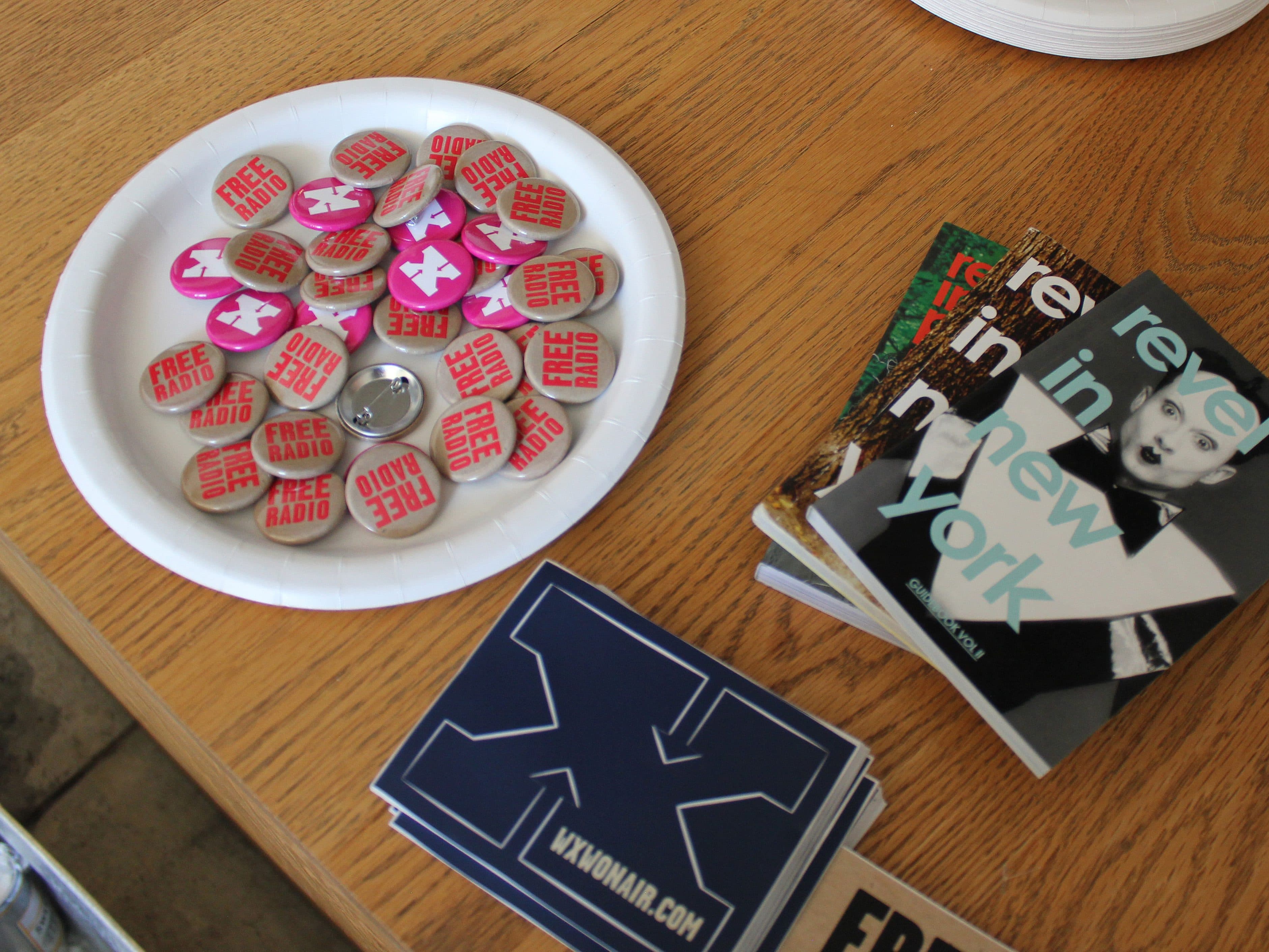 A table contains a paper plate filled with pink and red "Free Radio" buttons, a stack of stickers with a large blue letter "X,"  and three "revel in New York" booklets featuring a black and white photo of a person on the cover.