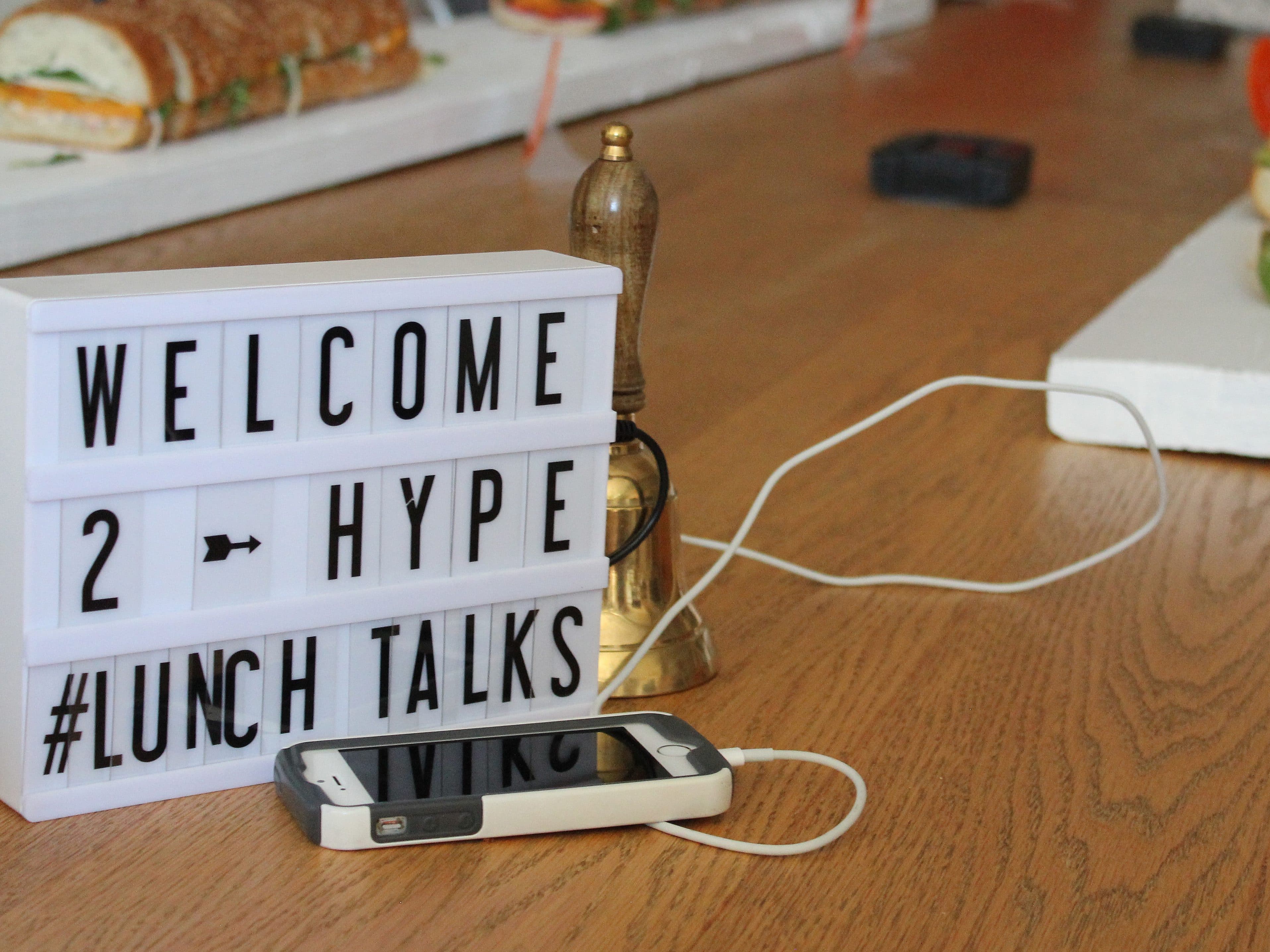 A lightbox sign on a table reads "WELCOME 2 HYPE #LUNCH TALKS." Beside it is a gold bell and a smartphone connected to a charging cable. Sub sandwiches are blurred in the background.