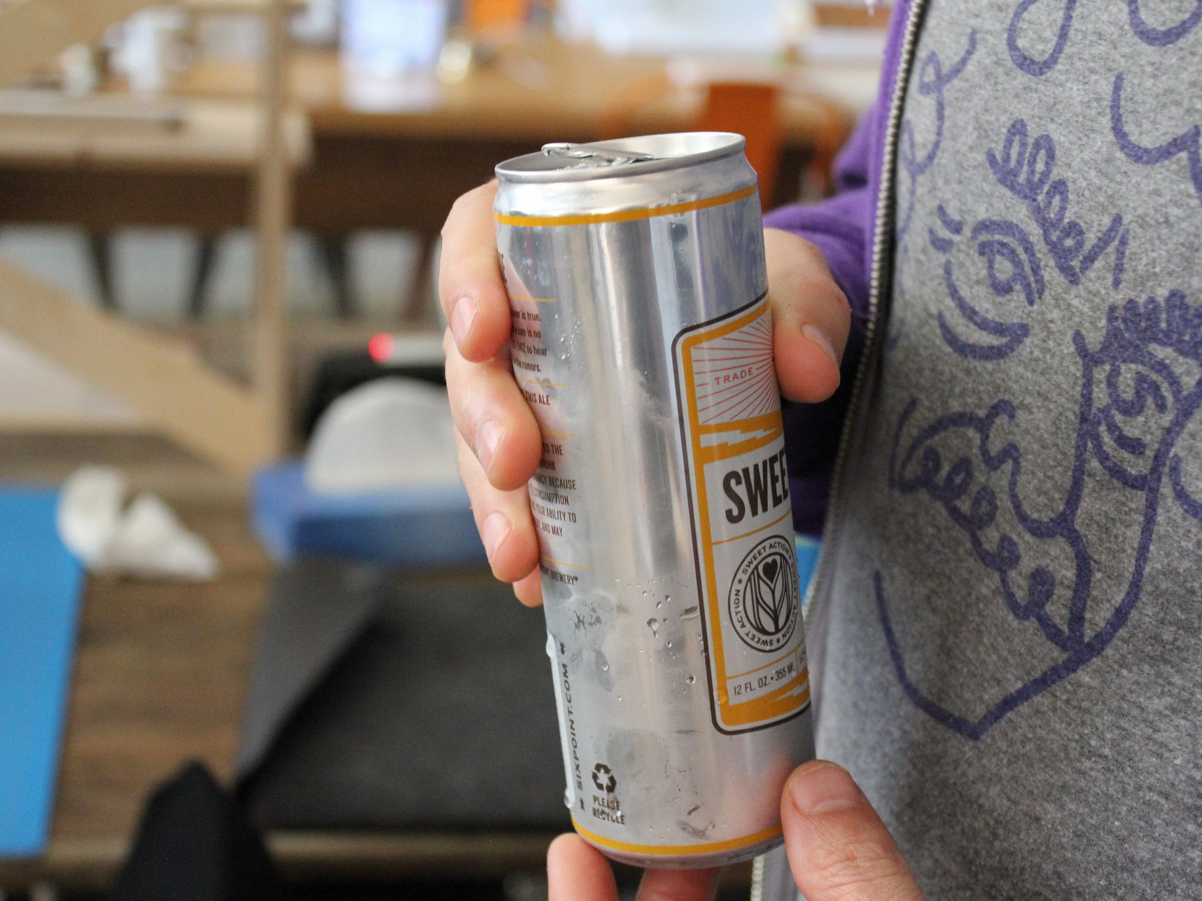 A person in a grey shirt with a purple swirl design is holding a silver can of "Sweet" beverage with an orange stripe near the top. In the background, there is a wooden table and some miscellaneous items.