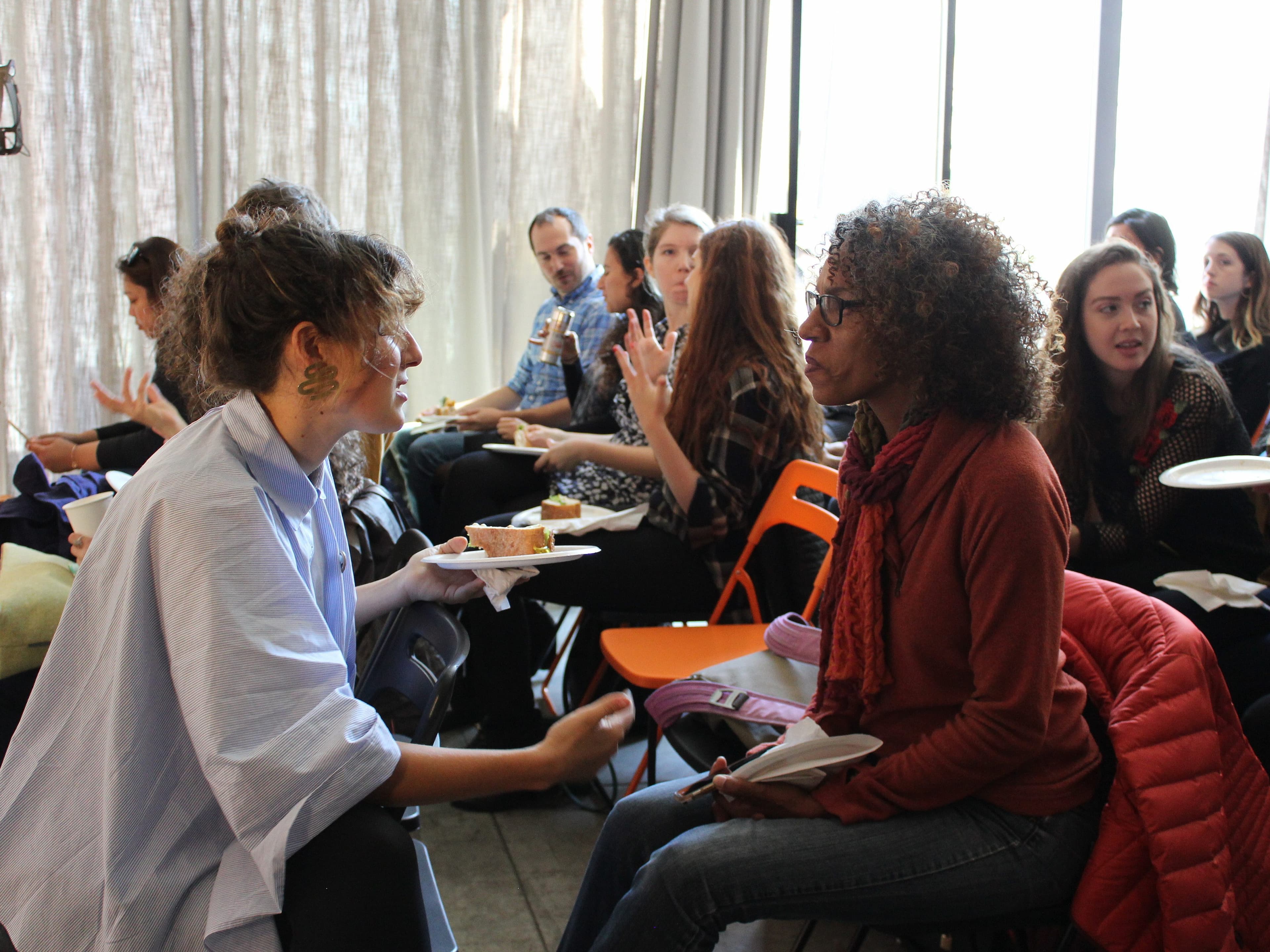 A group of people sit on orange and black chairs in a bright room with large windows. Two women in the foreground are seated, facing each other, and engaged in conversation while holding plates of food. Others in the background are also sitting and eating.