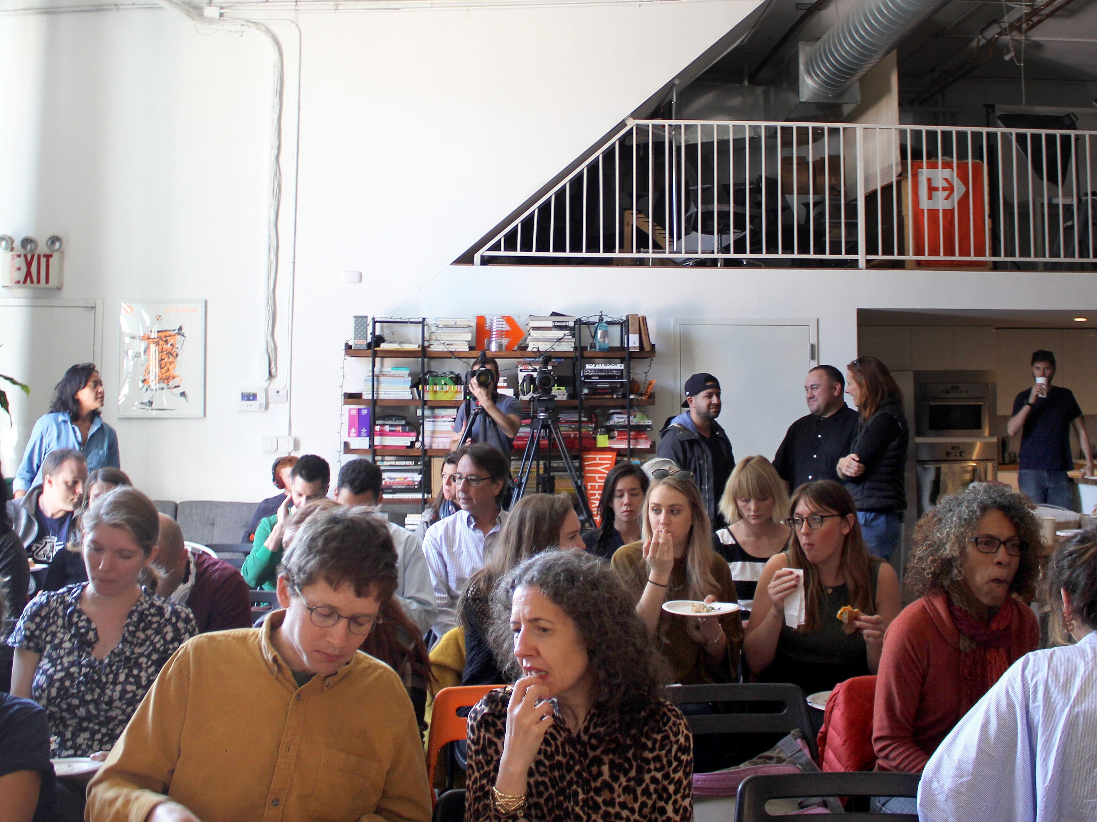 A group of people are sitting and standing in a well-lit room with an industrial design aesthetic. Some are eating, while others are chatting or looking at something off-camera. There is a loft above with a few people standing and observing the scene below.