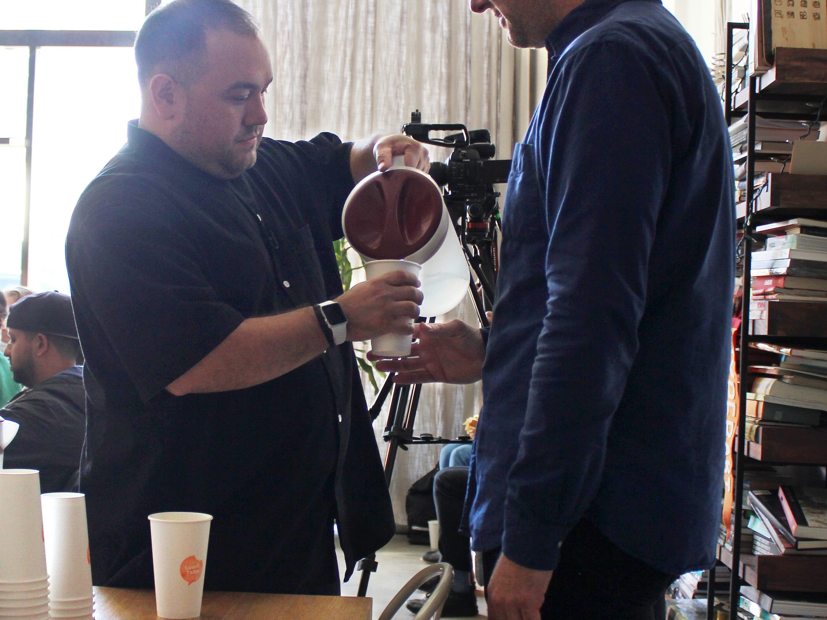 A man is pouring a dark beverage from a pitcher into a cup held by another man. They are standing beside a table with stacks of white paper cups and plates. Books and a camera can be seen in the background, with a well-lit room and large window.