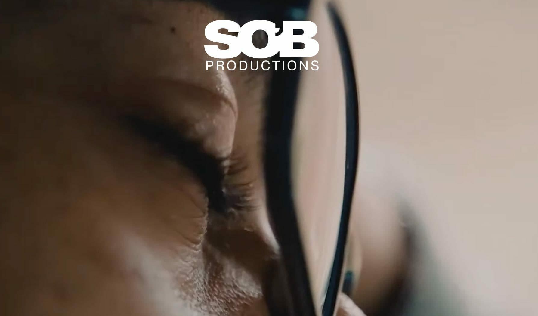 A close-up of a person's face with closed eyes, wearing black-framed glasses. The image has a soft focus, and "SOB Productions" is prominently displayed in white text at the top.