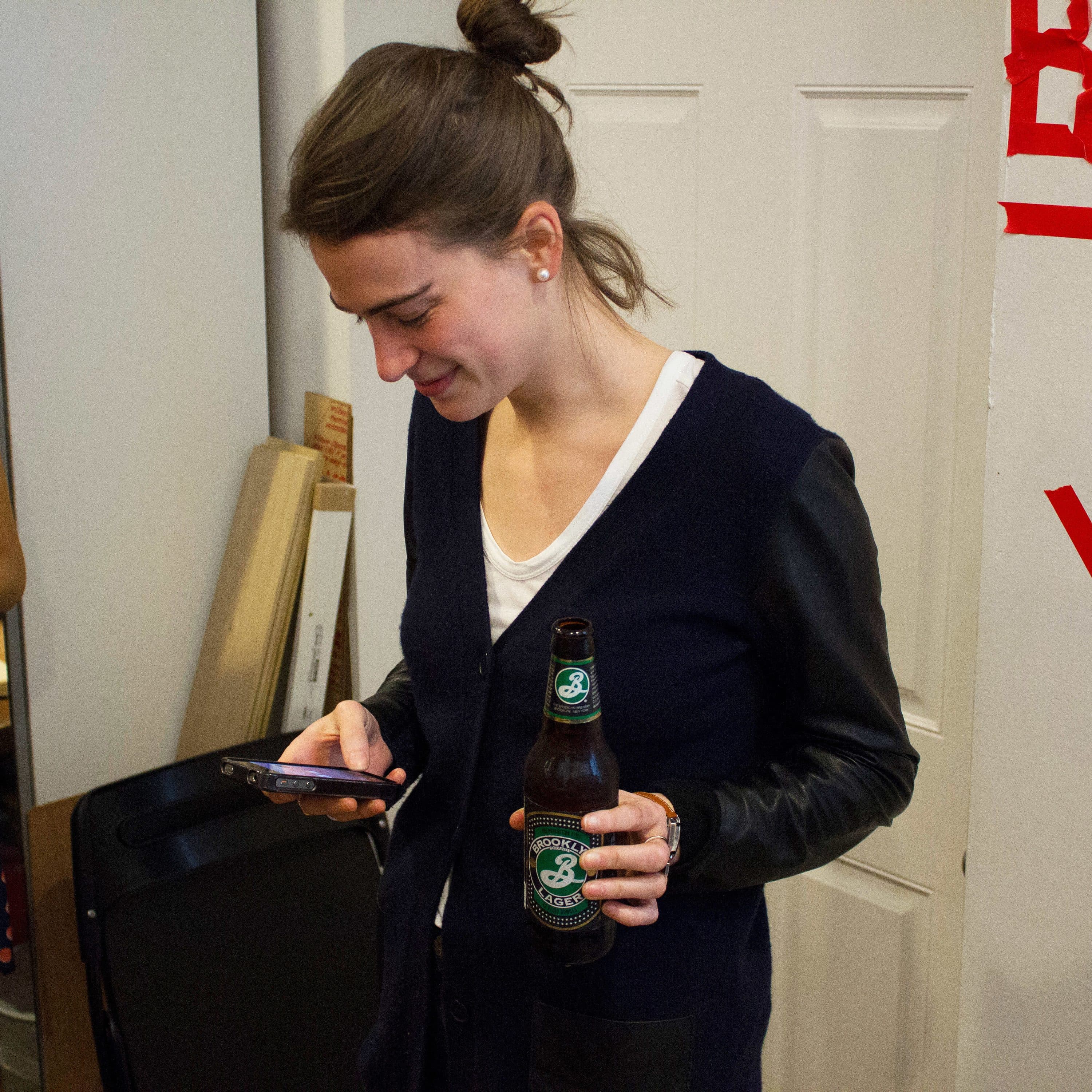 A woman with a bun hairstyle is holding a beer bottle in one hand and looking at her phone. She is wearing a cardigan. Another person in a white top and scarf is standing nearby, holding a plate with food. A wall with the word "BEER" and an arrow are visible.