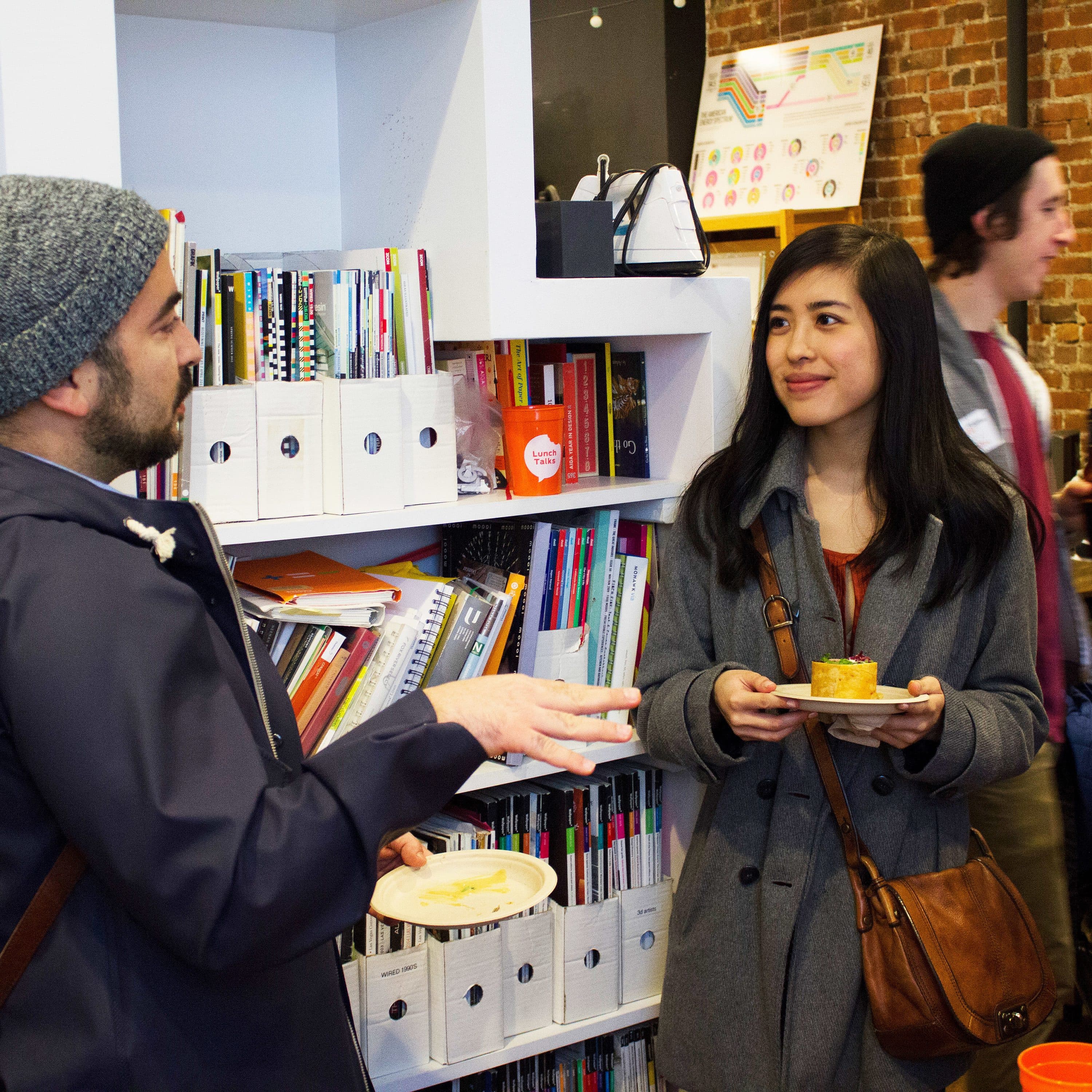 Two people, one in a beanie and backpack, and another in a gray coat, are conversing while holding plates with snacks in a room with bookshelves and colorful decor. More people are seen engaging in the background, suggesting a casual social gathering.