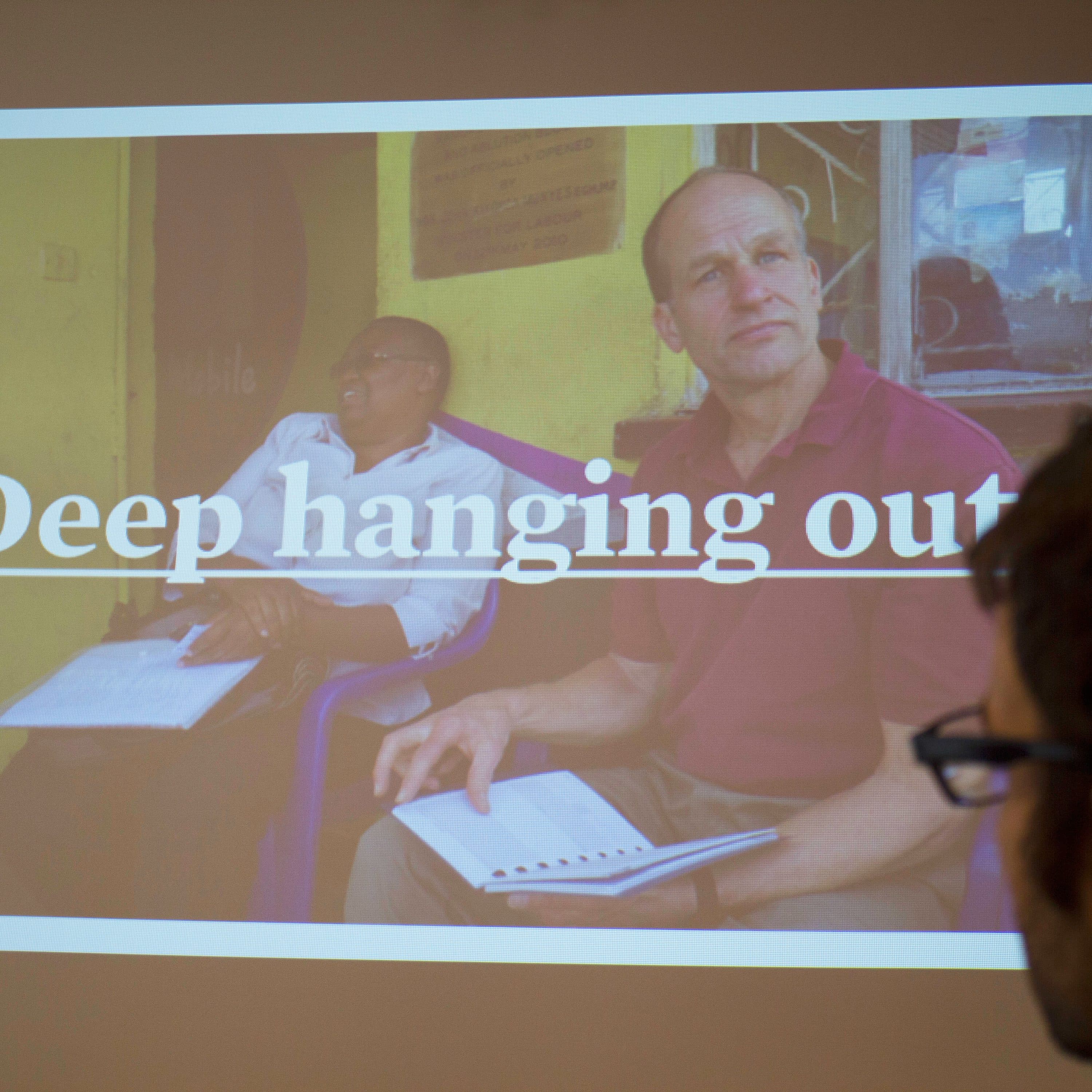 A man is in the foreground looking at a projected image on a screen. The image shows two men sitting outside, with one holding a notebook and the other relaxed. The text "Deep hanging out" is displayed prominently on the screen.