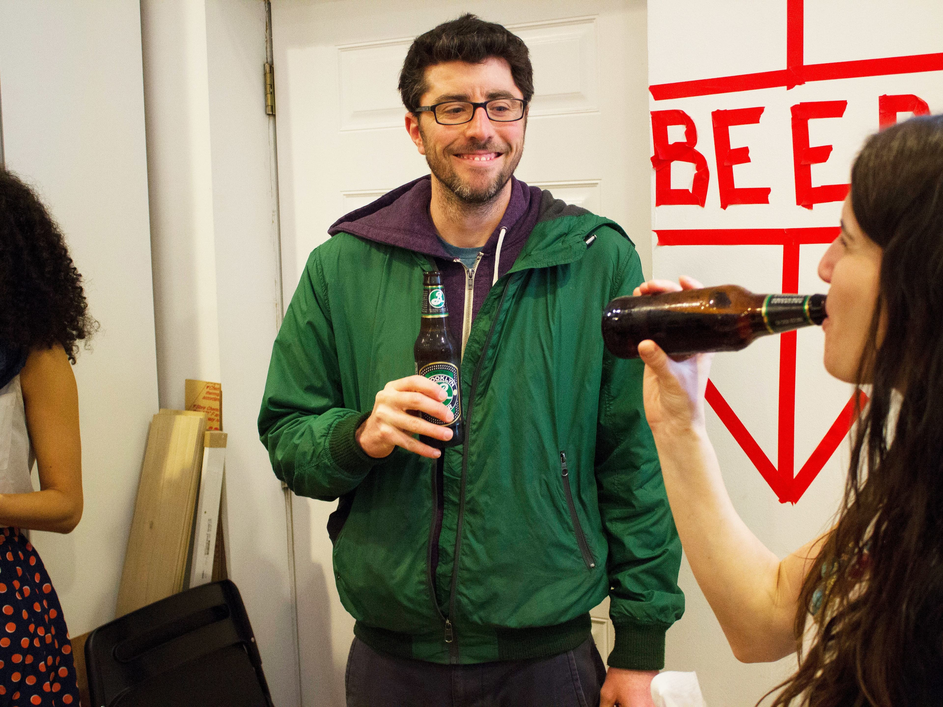 A man in a green jacket and glasses holds a bottle of beer and smiles while looking towards a woman drinking beer from a bottle. The woman has long hair and wears a patterned skirt. They are standing near a white wall with a red cross marked "BEER.