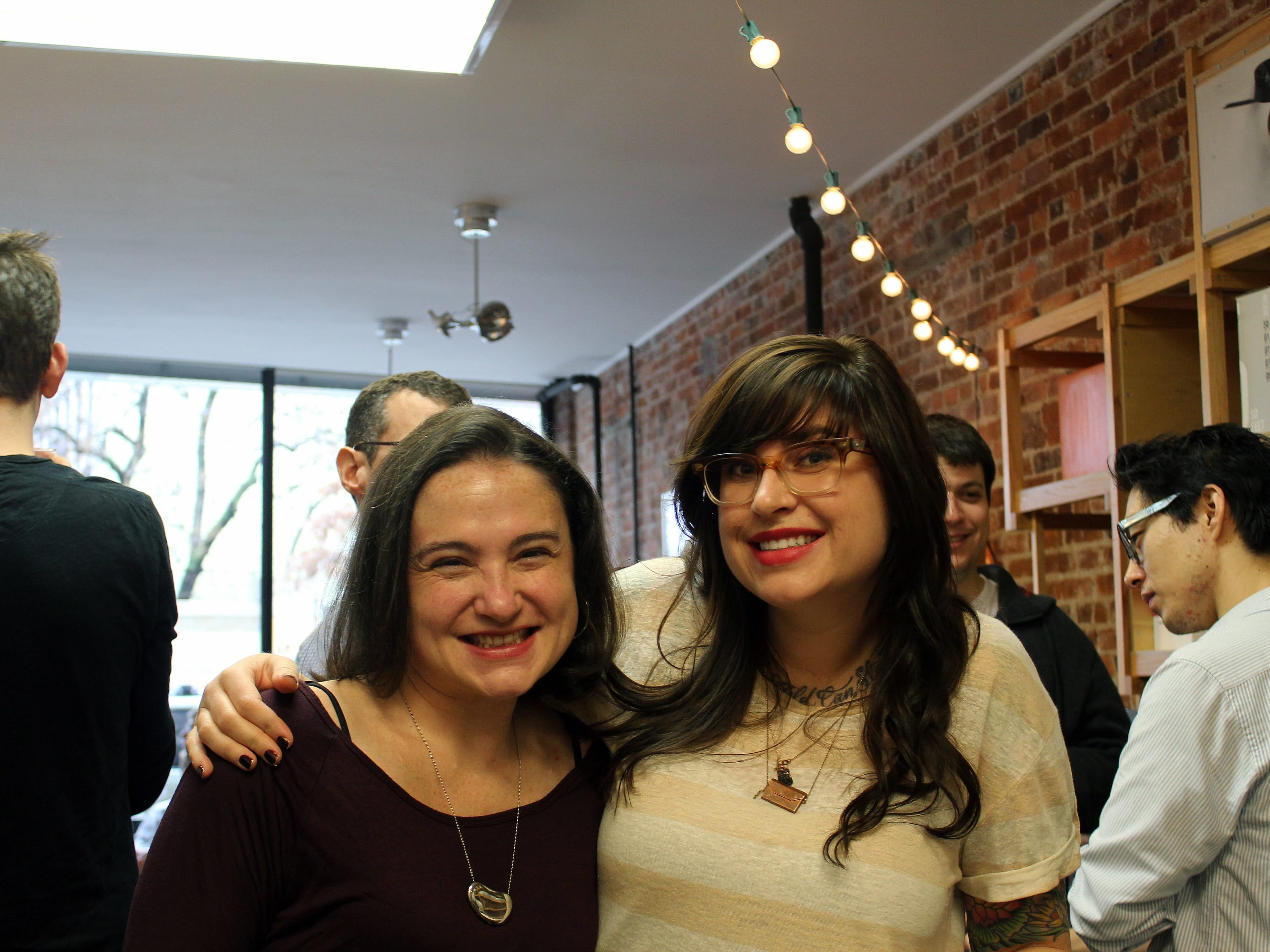 Two women are smiling at the camera with their arms around each other in a room with exposed brick walls. Other people are seen in the background, engaging in conversation. The ceiling has string lights hanging down, and there's a large window letting in natural light.