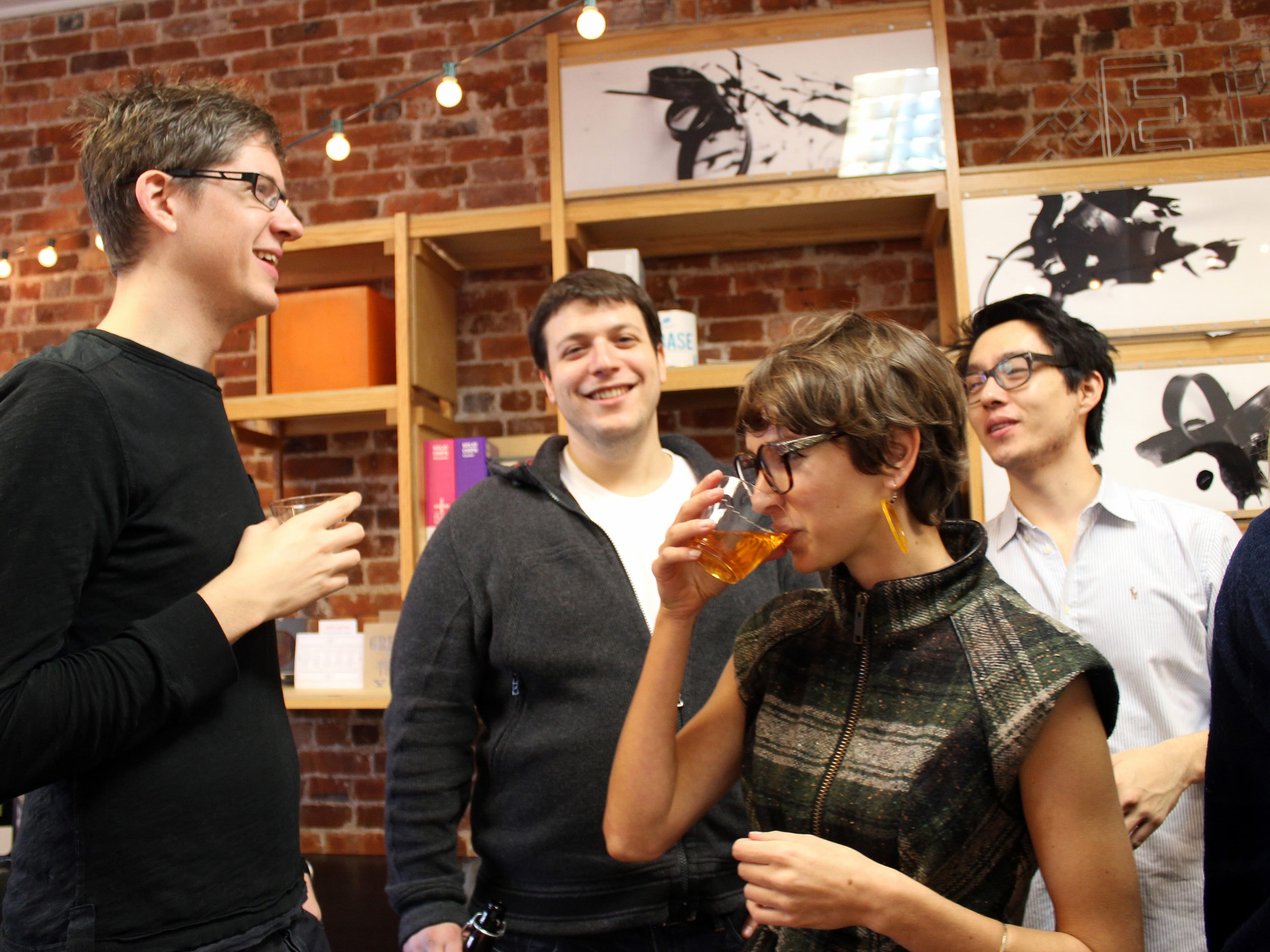 A group of four people are socializing in an indoor space with brick walls. One person is drinking from a glass, while the others are engaged in conversation. Shelves with various items and art hang on the wall behind them, and string lights add a cozy ambiance.