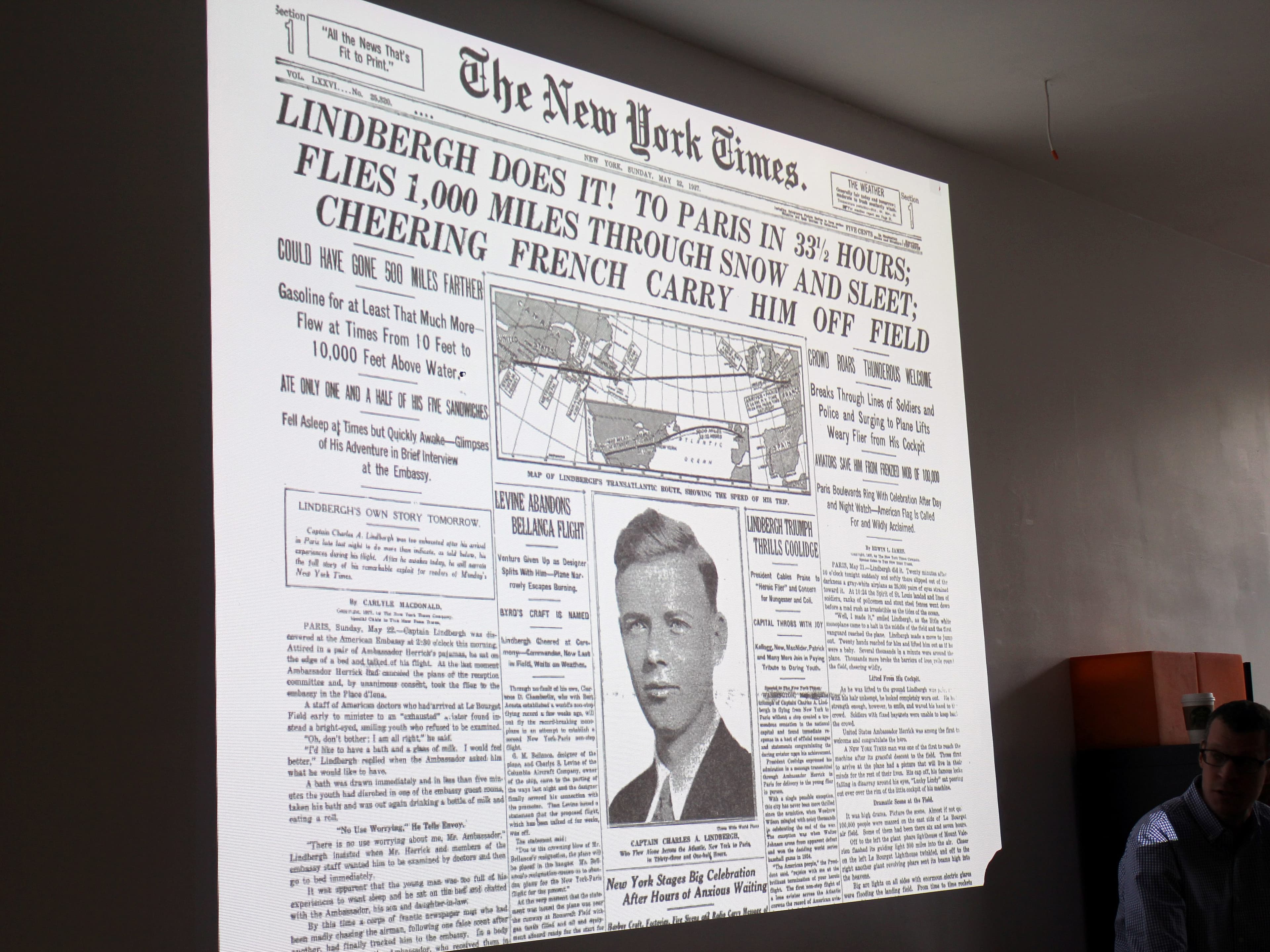 A projected image of a historical New York Times front page featuring Charles Lindbergh's transatlantic flight. The headline reads "Lindbergh Does It! To Paris in 33½ Hours". A man is partially visible on the right side of the picture.