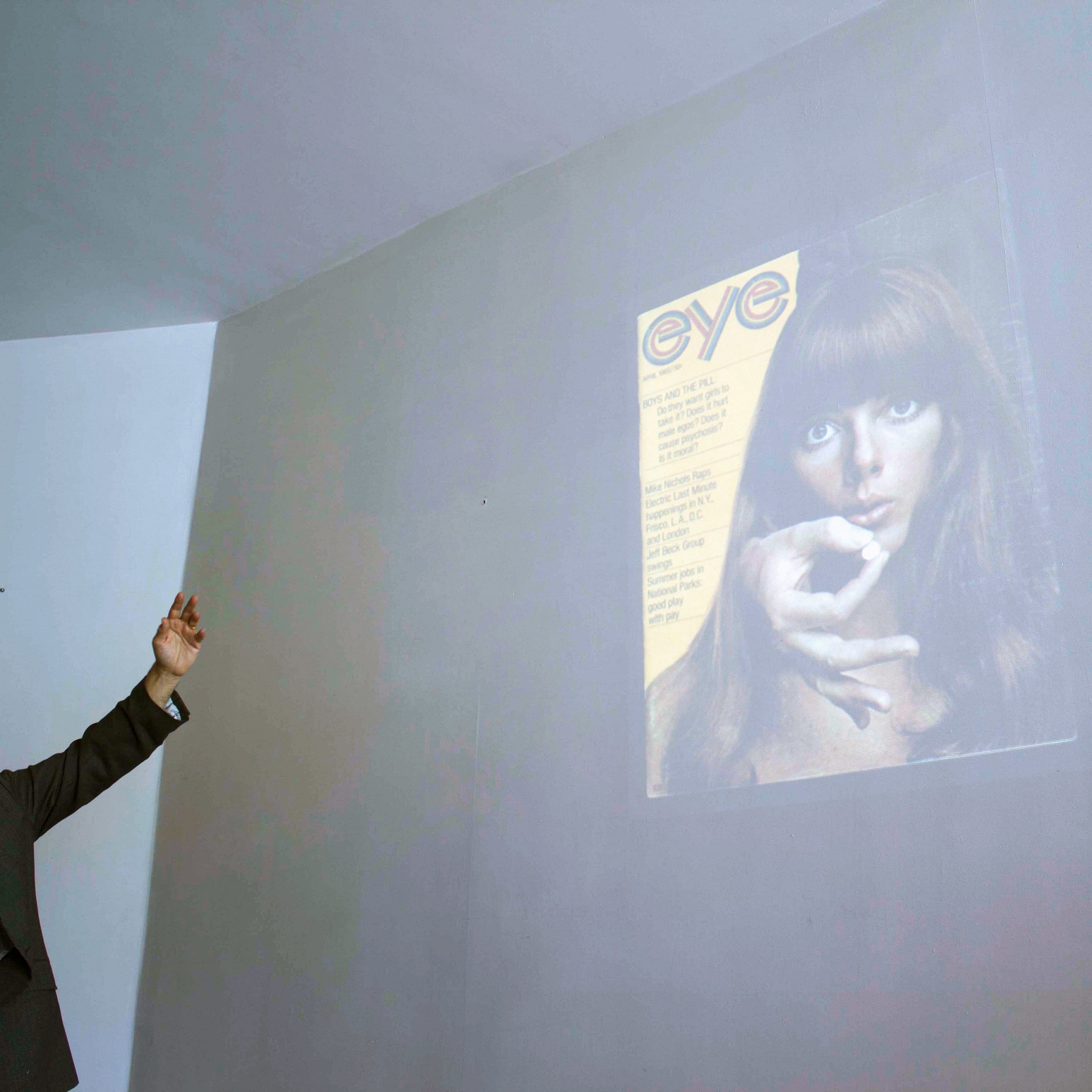 A man stands pointing towards a wall where an old magazine cover is projected. The magazine, titled "eye," features a woman with long hair holding a finger to her lips. The man holds a microphone and appears to be giving a presentation.