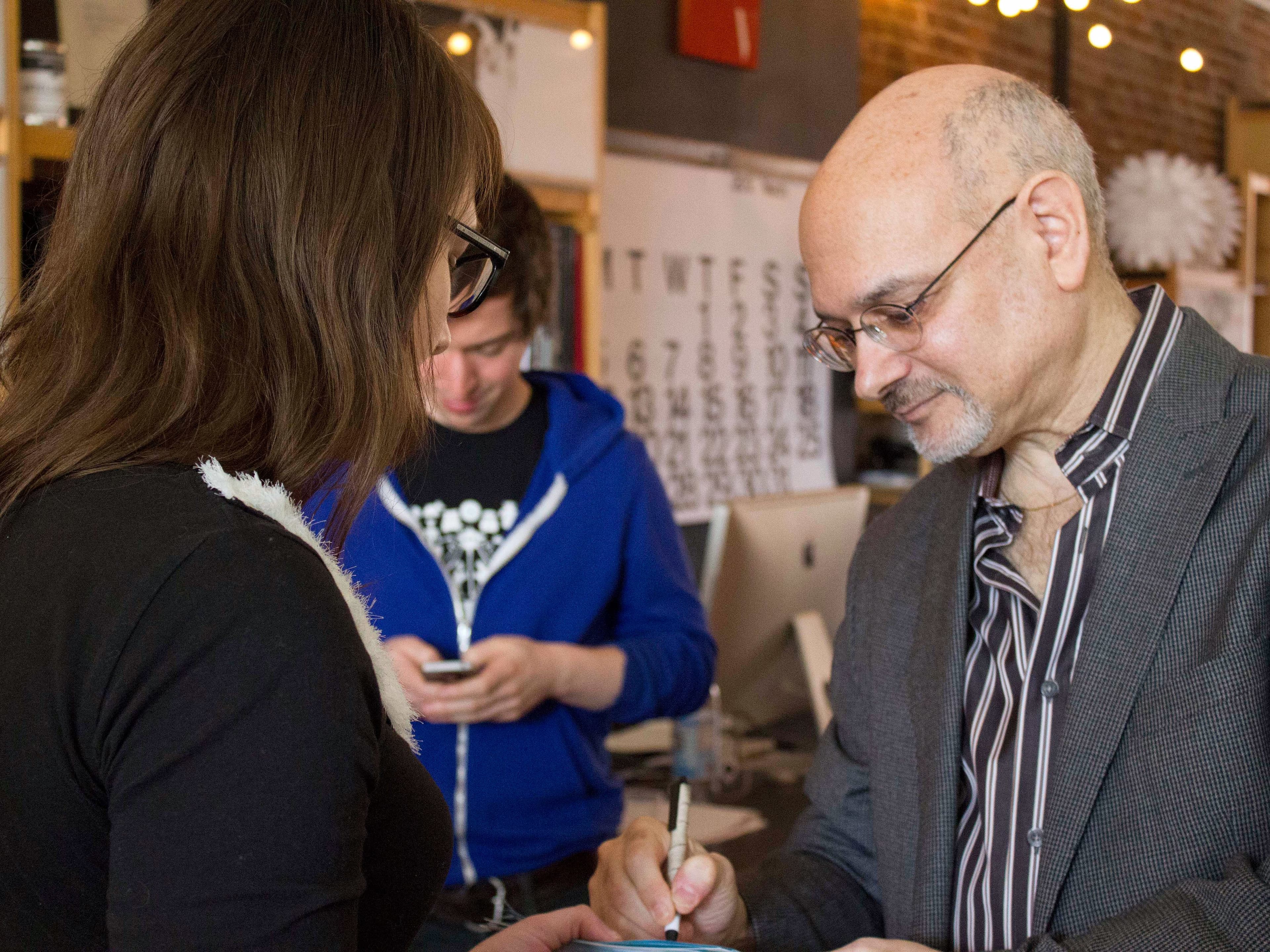 A man with glasses and a partially bald head is signing something for a woman. He is wearing a striped shirt and a blazer. The woman, with long brown hair, faces him. In the background, a younger man in a blue hoodie is looking down, using his phone.