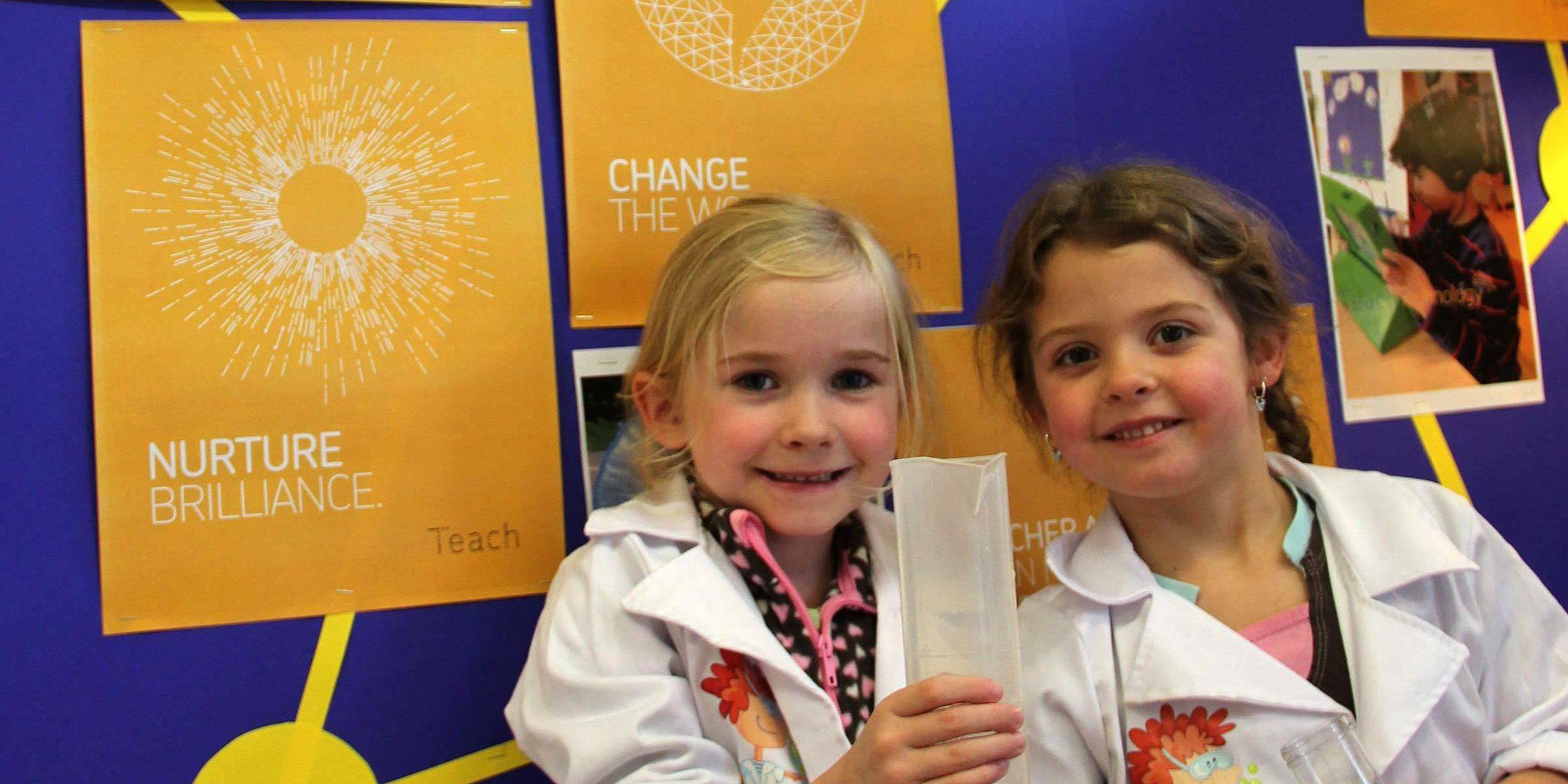 Two young girls wear scientist lab coats while smiling and holding a paper tube in a classroom. Behind them, colorful posters display phrases like "Nurture Brilliance" and "Change the World." The setting appears vibrant and educational with a focus on learning.