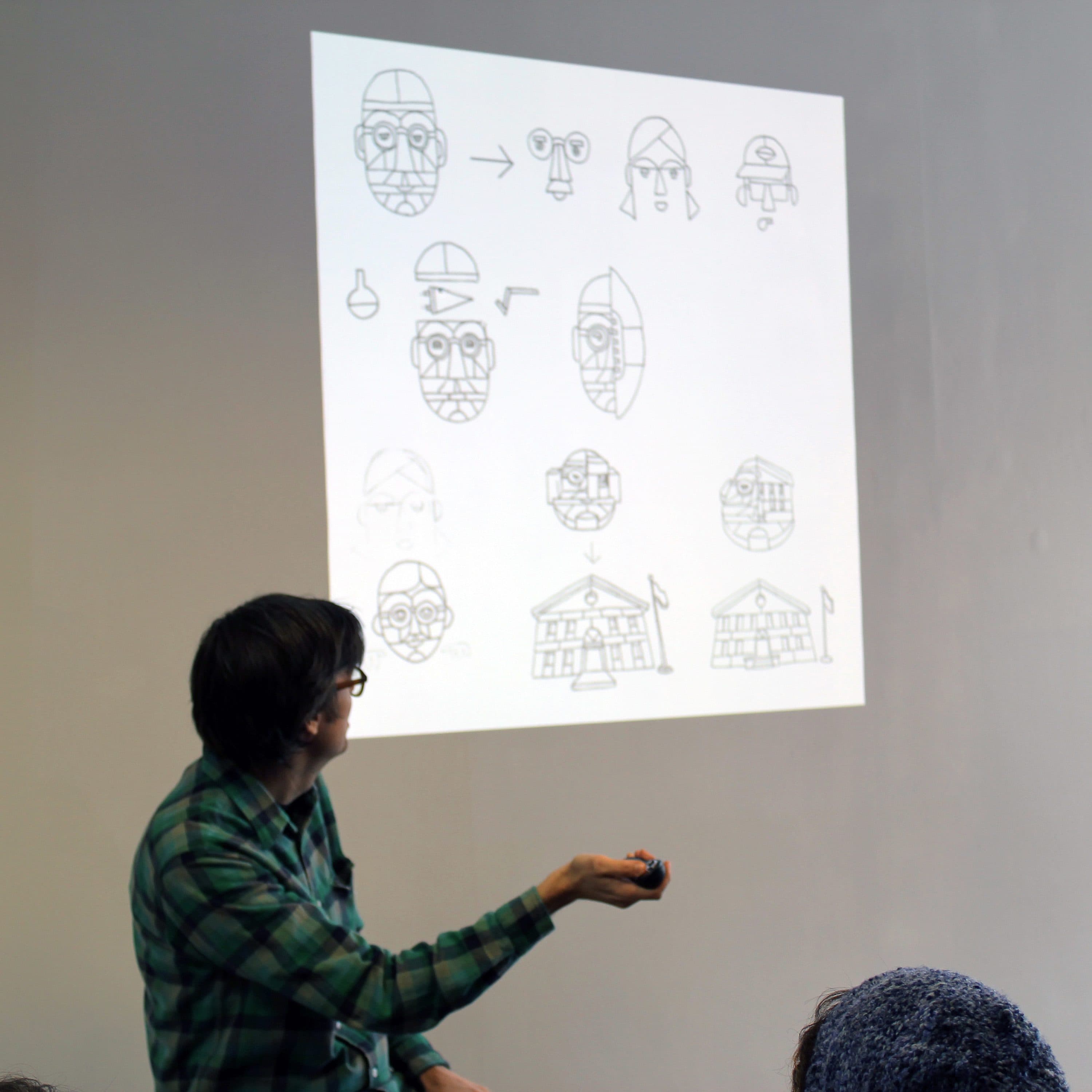 A person in a green plaid shirt is giving a presentation. They point towards a projection on the wall which displays a series of sketches showing the progression of design elements for a character or object. The audience faces the presenter.