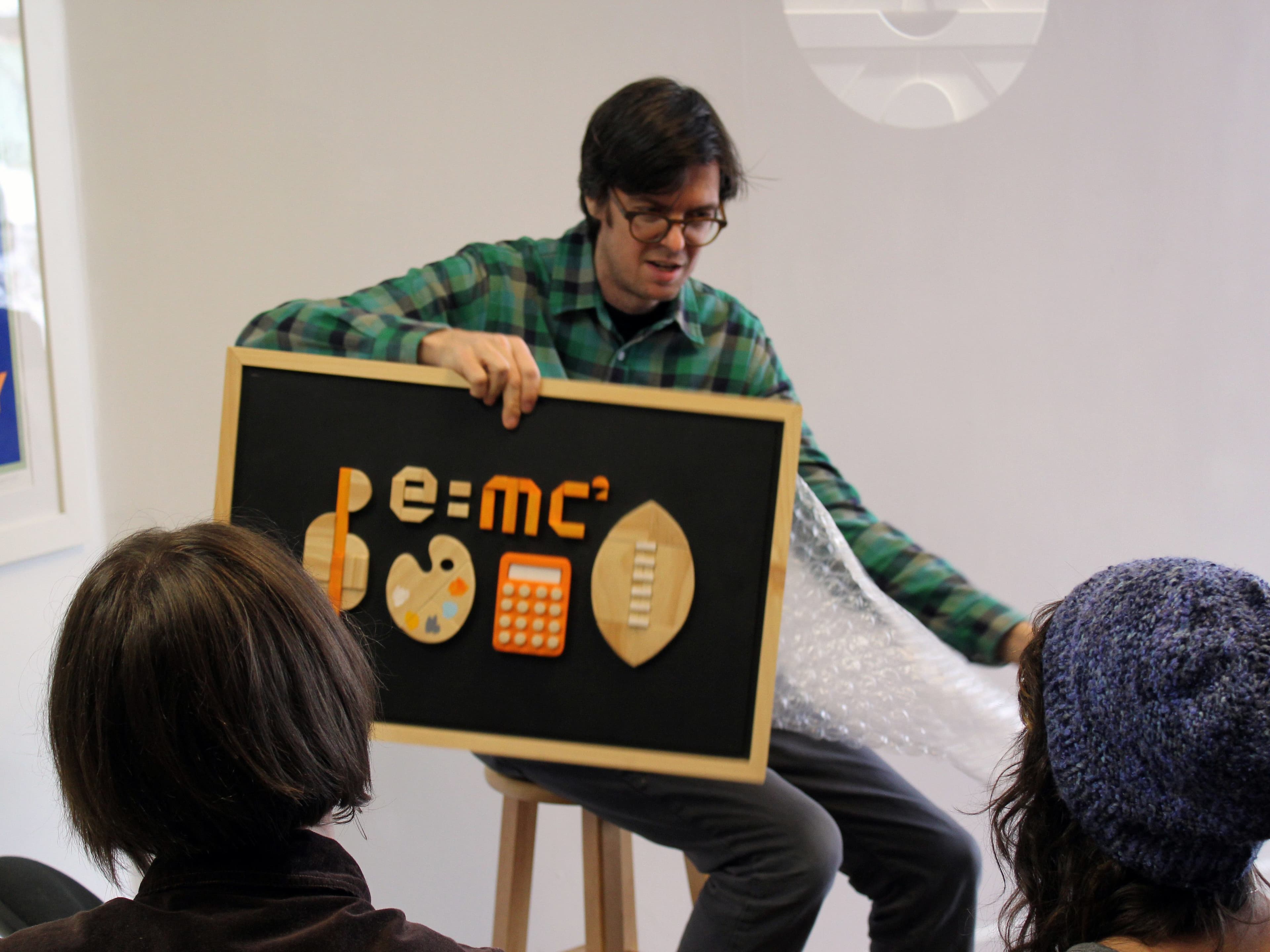 A person in a green plaid shirt sits on a stool and holds a sign with various mathematical symbols, including E=mc². They are talking to an audience, with three people visibly listening, two with their backs to the camera, one partially seen in front.