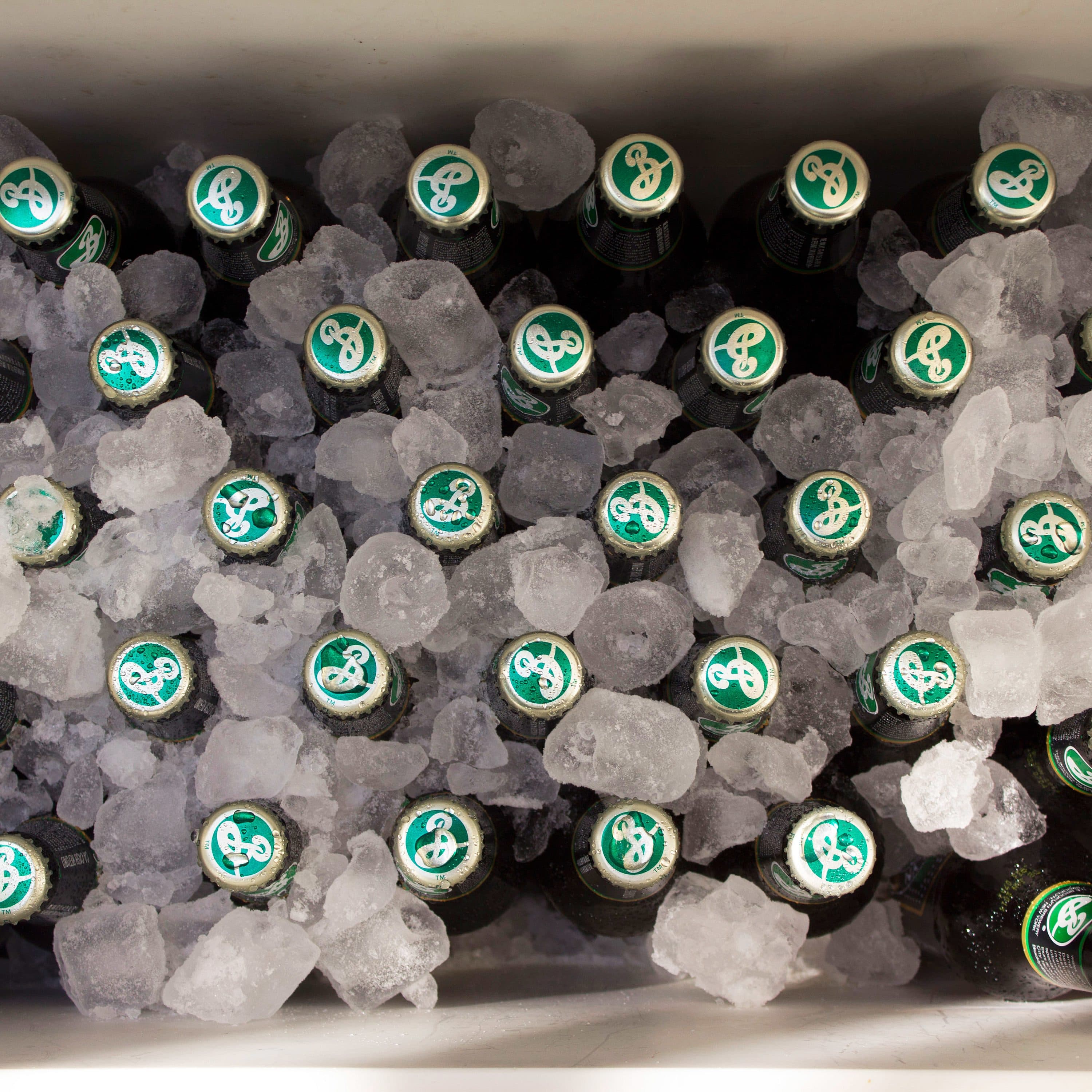A cooler filled with ice and several green-capped bottled beverages lying on their sides. The bottles have a logo with a stylized "B" on the caps. The ice is evenly distributed, keeping the bottles cold.