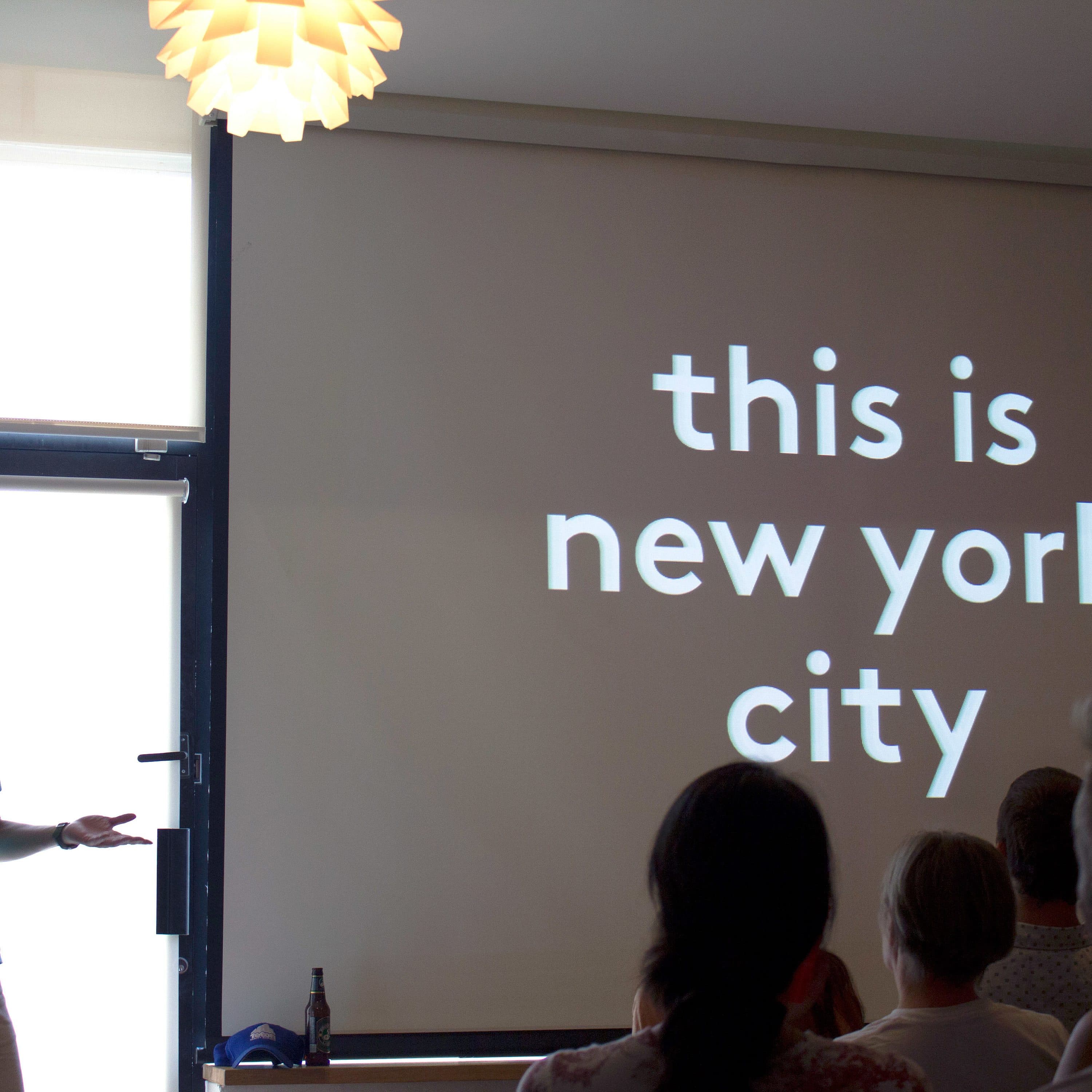 A person stands in front of an audience, presenting a slide that reads "this is new york city." The presentation is indoors with modern lighting fixtures visible on the ceiling. The person appears to be gesturing while speaking, holding a remote in one hand.