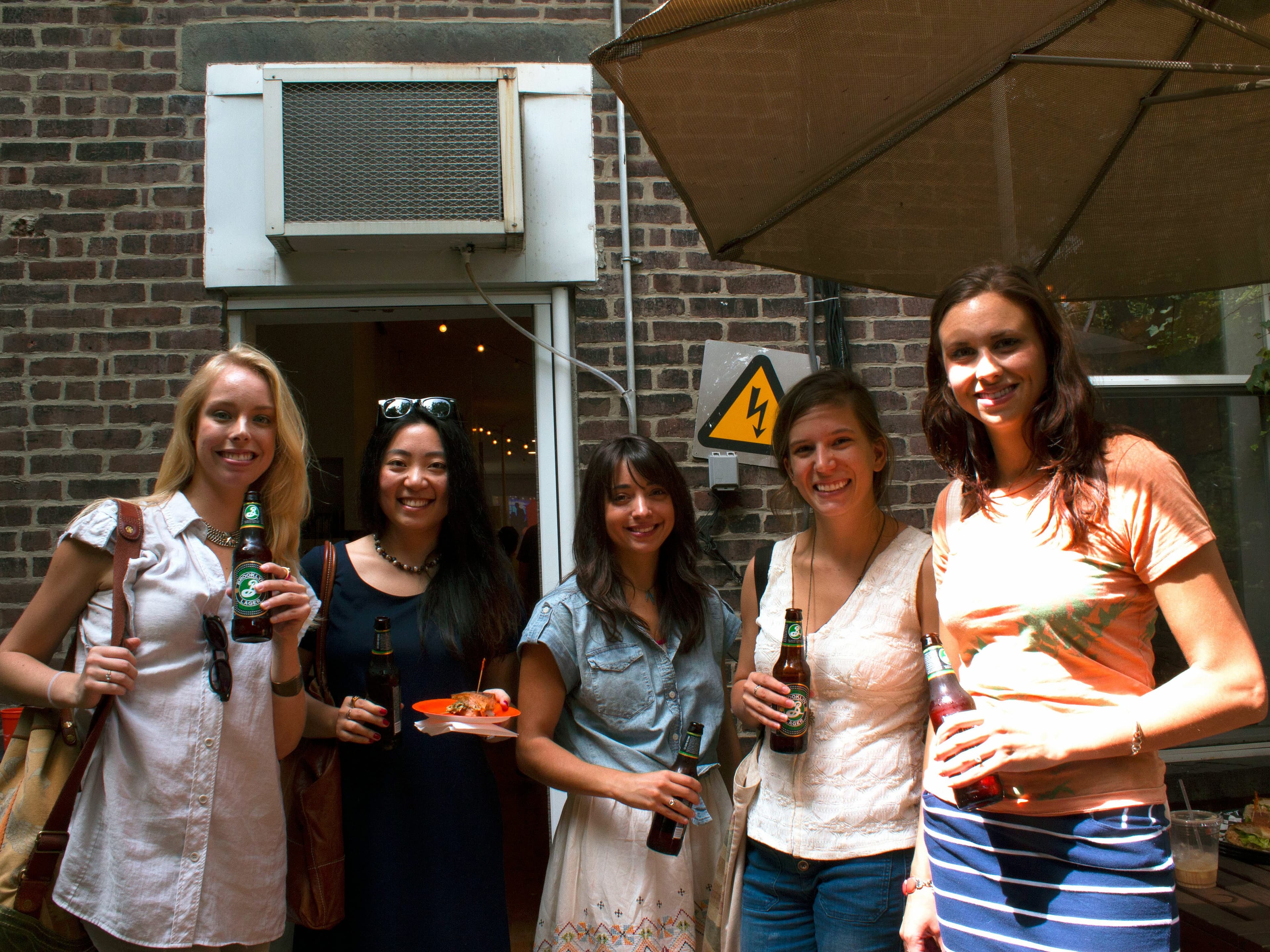 Five women are standing together under an umbrella outside, holding drinks and smiling at the camera. One of them holds a plate of food. A brick wall with an air conditioning unit and a yellow caution sign is in the background.