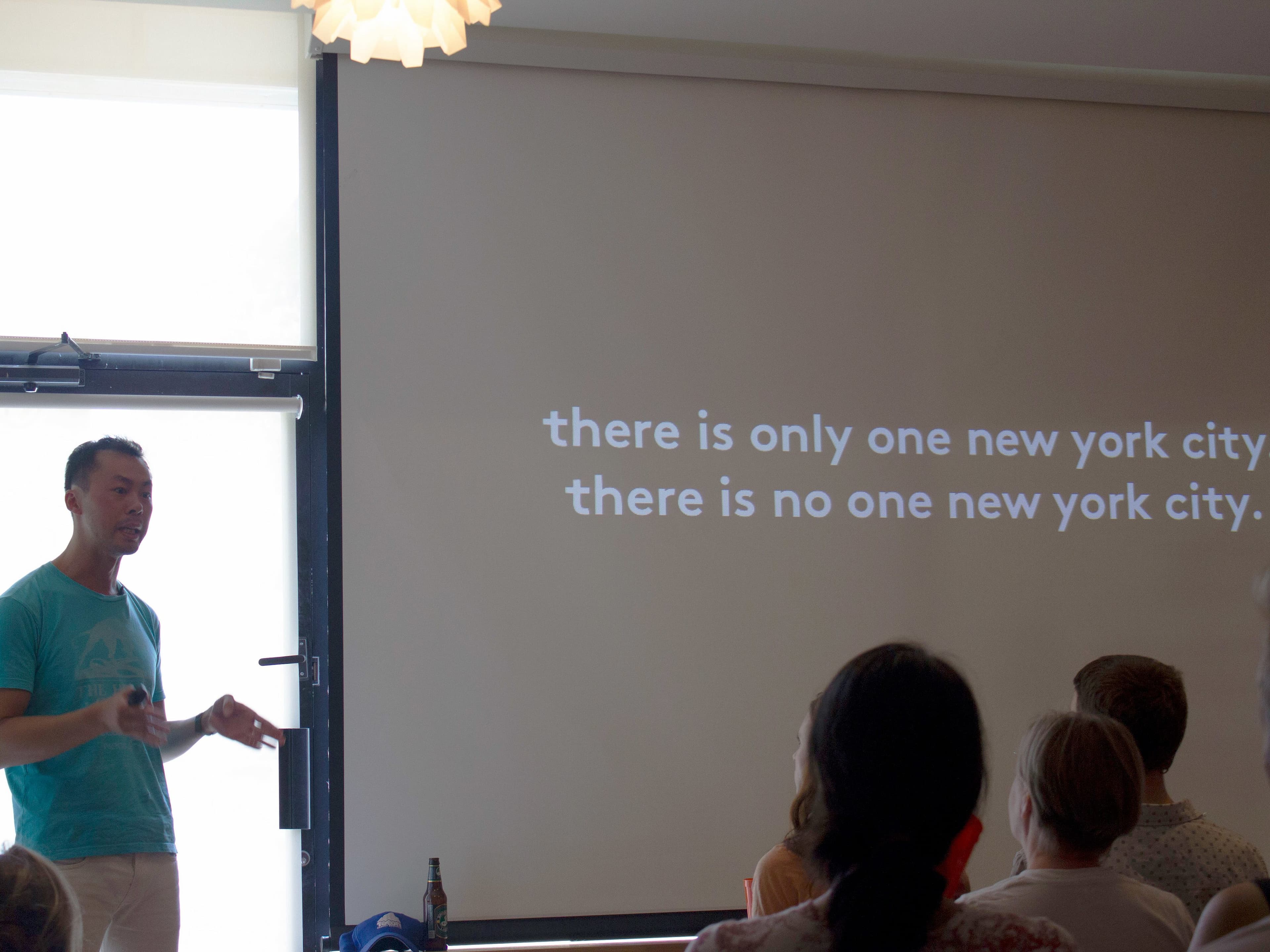 A man in a turquoise shirt is giving a presentation in front of an audience. Behind him, a projection screen displays the text: "there is only one new york city. there is no one new york city." The room is dimly lit, and a window is visible on the left.