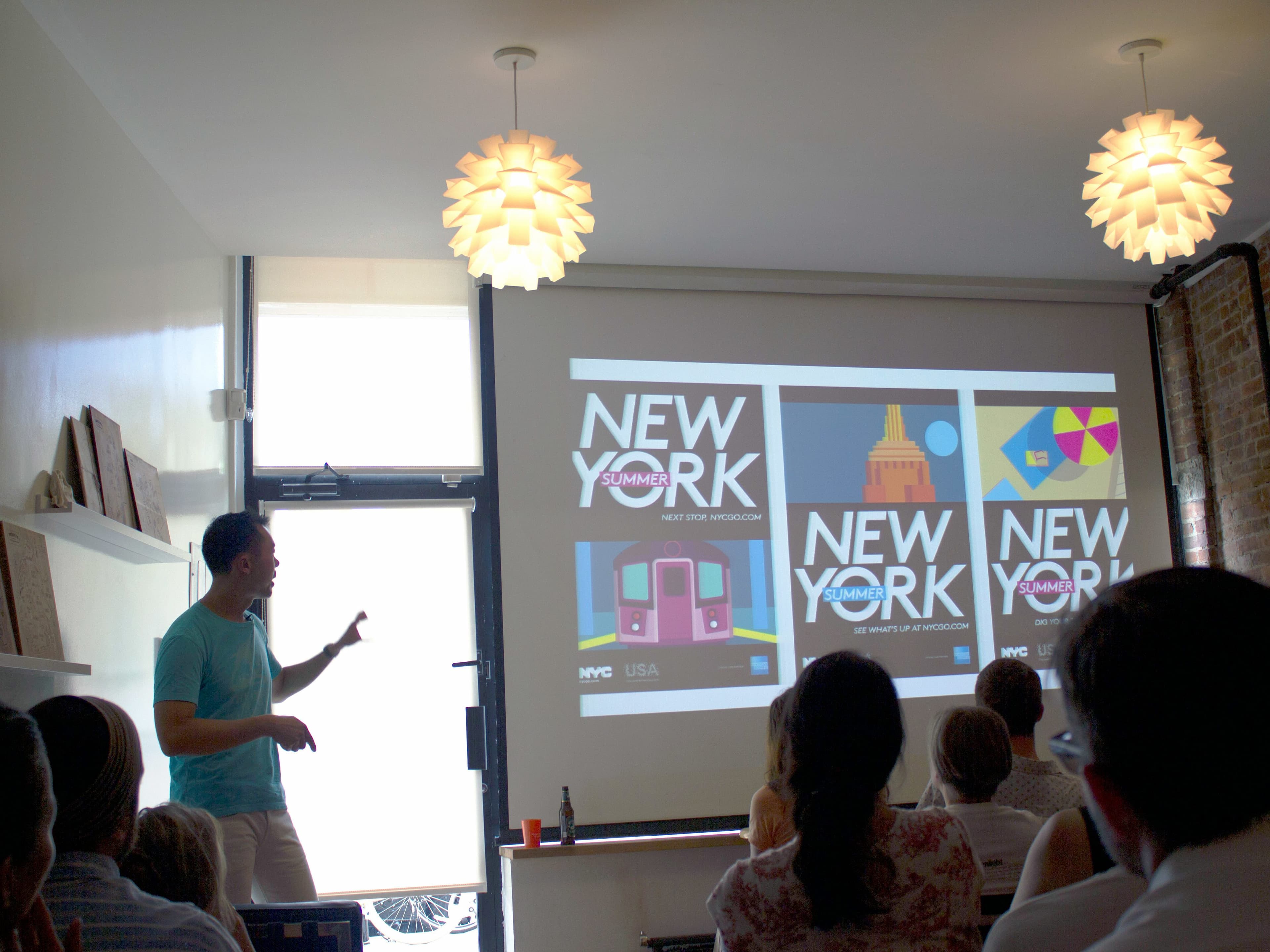A person is giving a presentation in a dimly lit room, pointing to a large screen displaying four slides with "NEW YORK" in bold text. An audience seated in front is watching attentively. The room has modern hanging lights and an exposed brick wall.