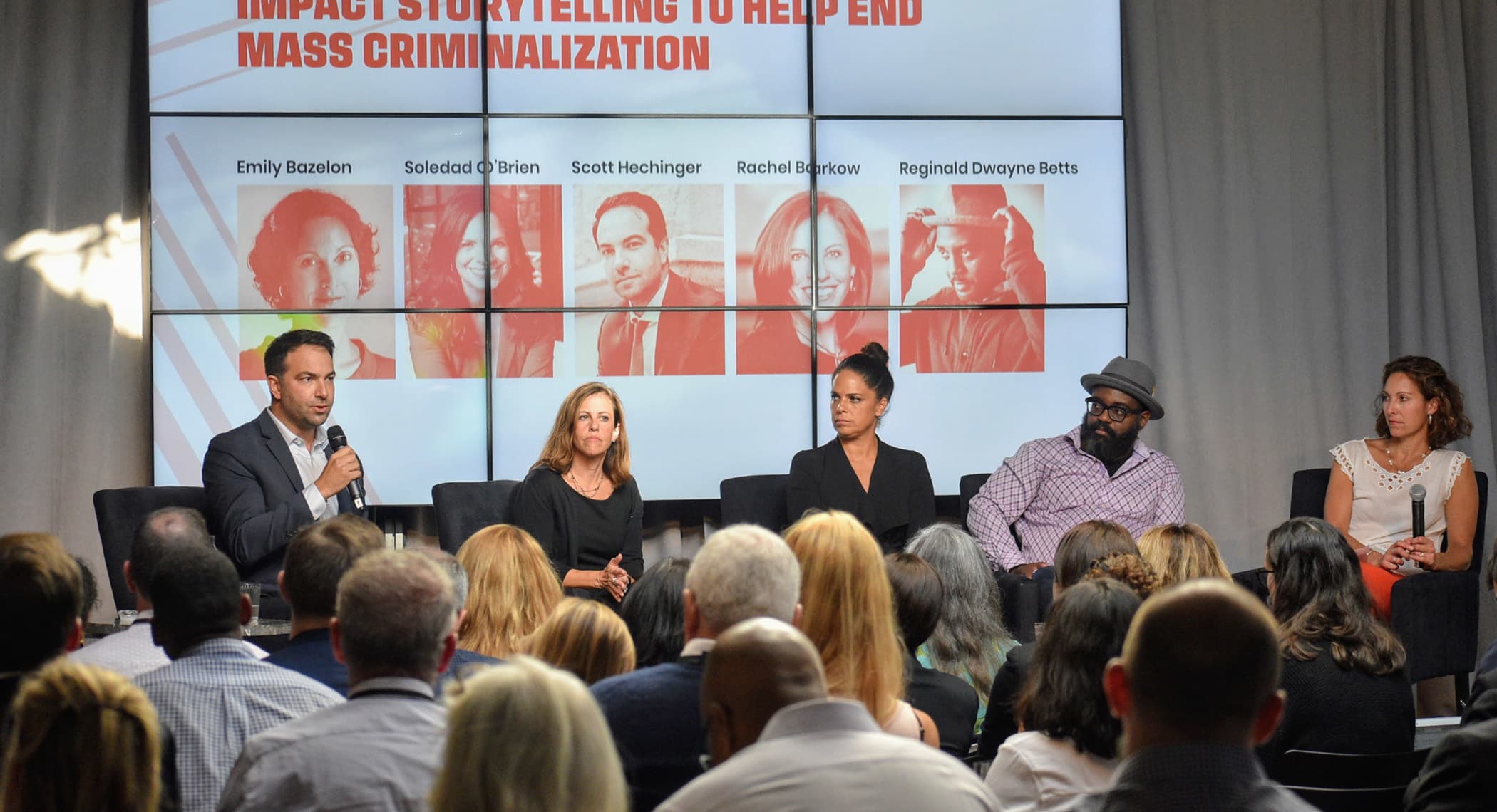 A panel discussion with five speakers seated on stage in front of an audience. Behind them is a screen displaying "IMPACT STORYTELLING TO HELP END MASS CRIMINALIZATION" with names and photos of the speakers: Emily Bazelon, Soledad O'Brien, Scott Hechinger, Rachael Rollins, Reginald Dwayne Betts.