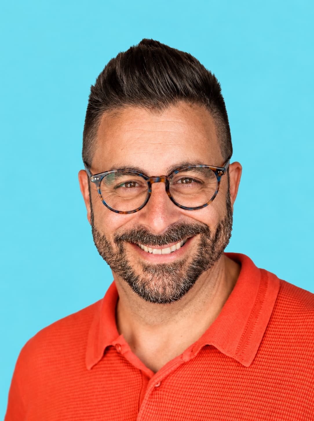Smiling man with short dark hair, beard, and glasses wears an orange collared shirt, posing in front of a plain light blue background.