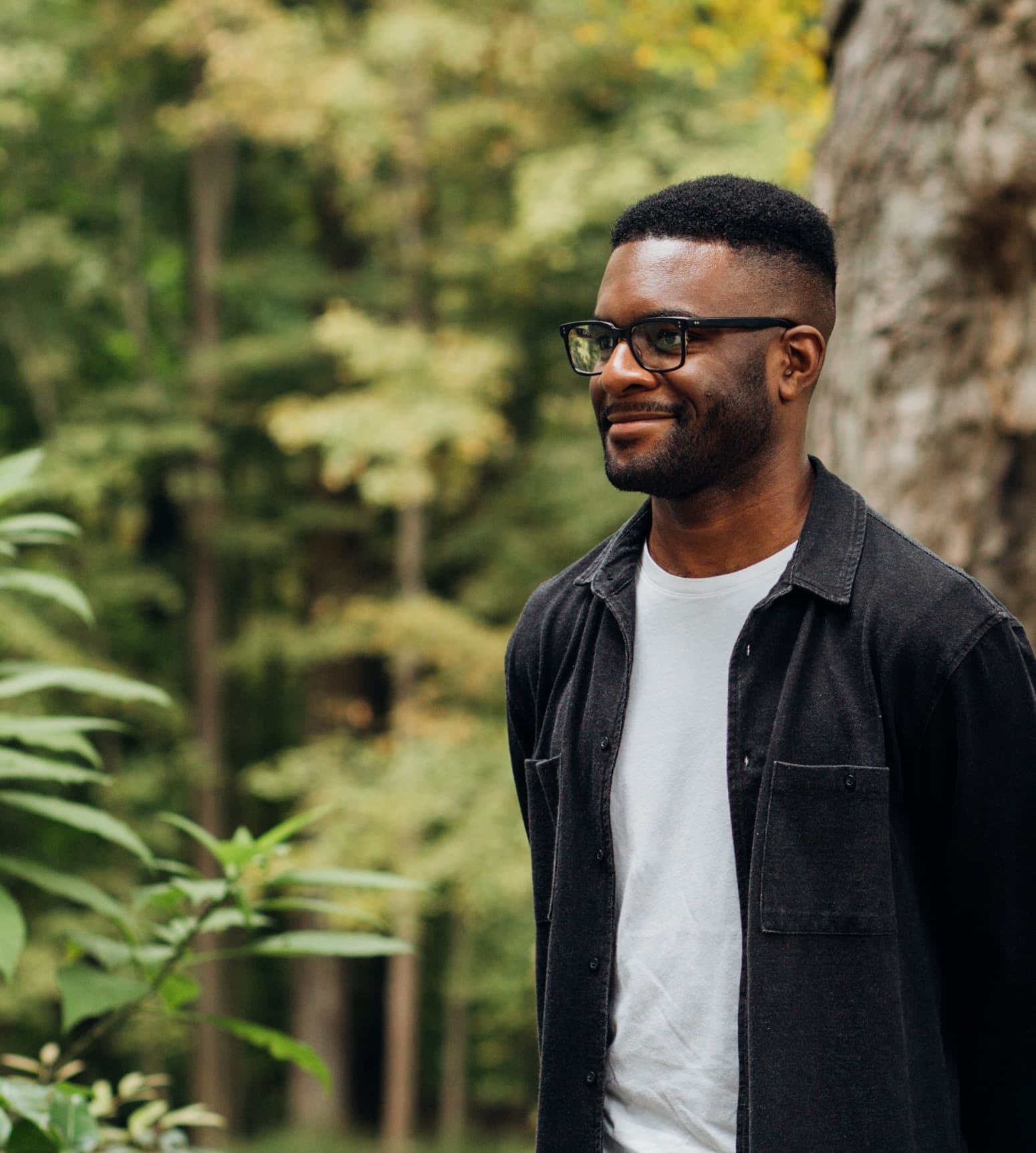A man wearing glasses, a white t-shirt, and a dark button-up shirt stands outdoors in a forested area, smiling and looking to the right. The background features green trees and foliage, highlighting the natural setting.