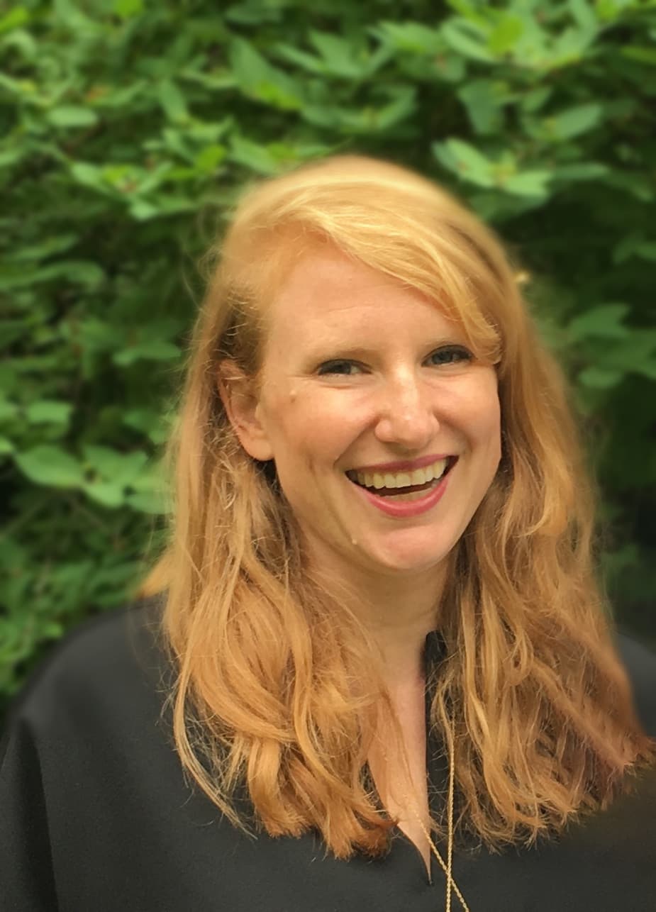 A woman with long, wavy red hair is smiling brightly at the camera. She is wearing a dark blouse and is standing outdoors with a backdrop of green foliage.