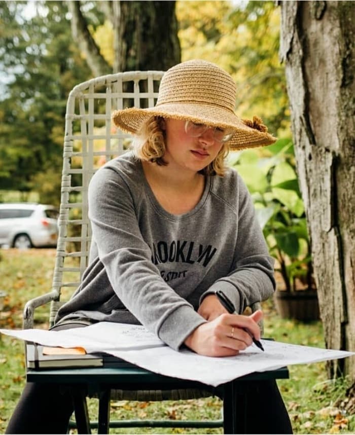 A person with light-colored hair is sitting on a chair outdoors, wearing a wide-brimmed straw hat and a gray "Brooklyn" sweatshirt. They're intently writing or drawing on a sketch pad placed on a small table. Some trees and a parked car can be seen in the background.