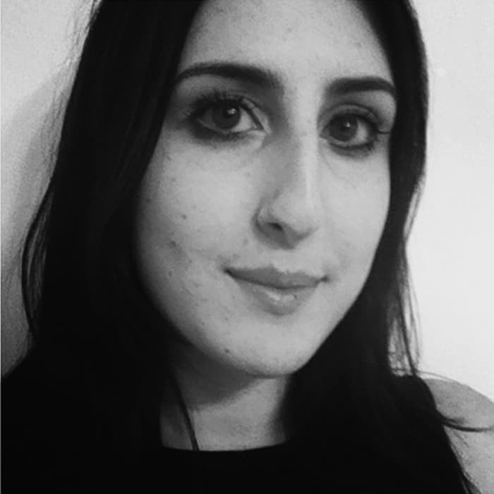 Black and white close-up photo of a person with long, dark hair and dark eyes, slightly smiling, looking directly at the camera. They have a light complexion and are wearing a dark top. The background is plain and light-colored.