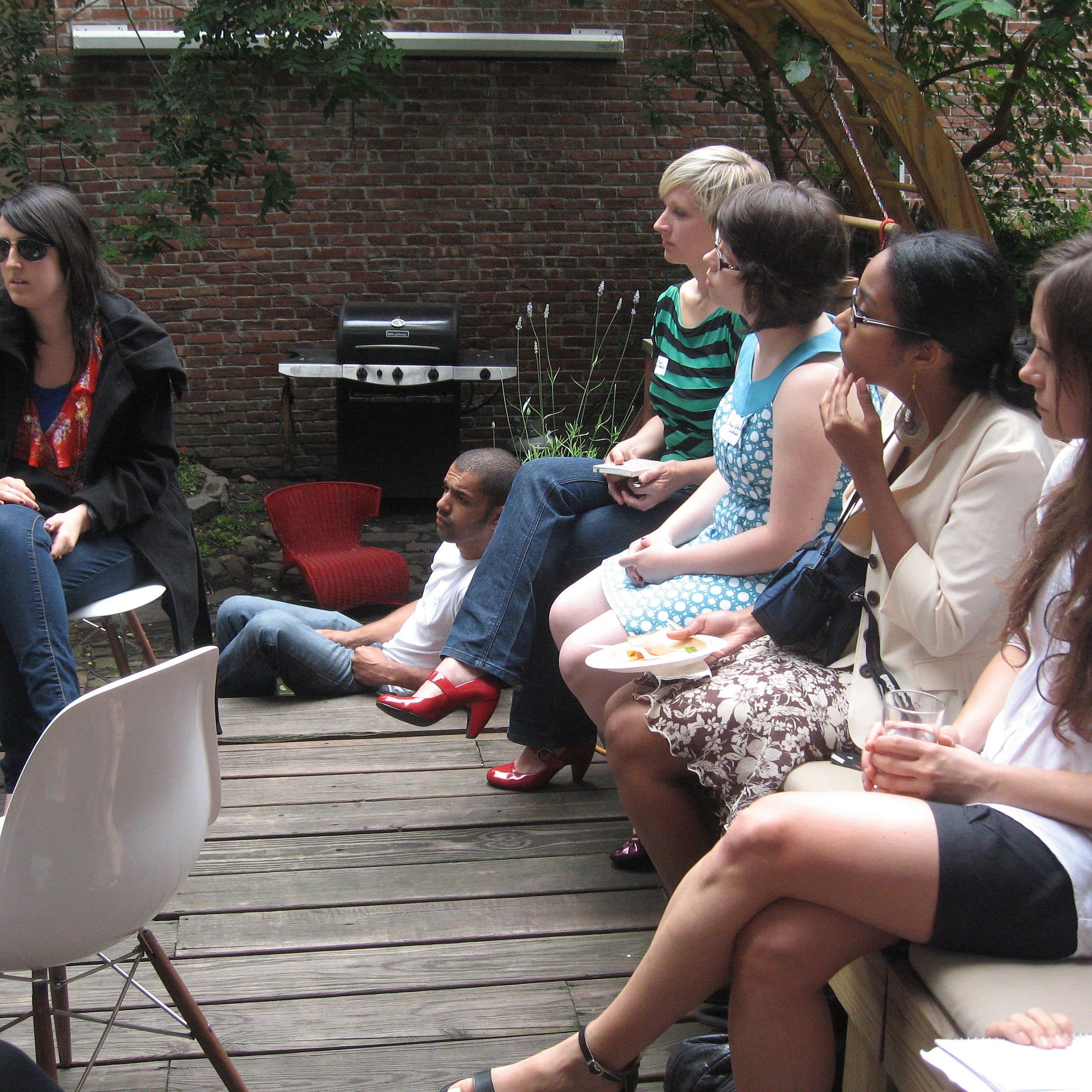 A group of people sit and stand on a wooden deck during an outdoor gathering. Some are seated on chairs while others sit on the ground. A woman with red shoes stands out. Brick walls and greenery frame the background. The atmosphere appears casual and social.