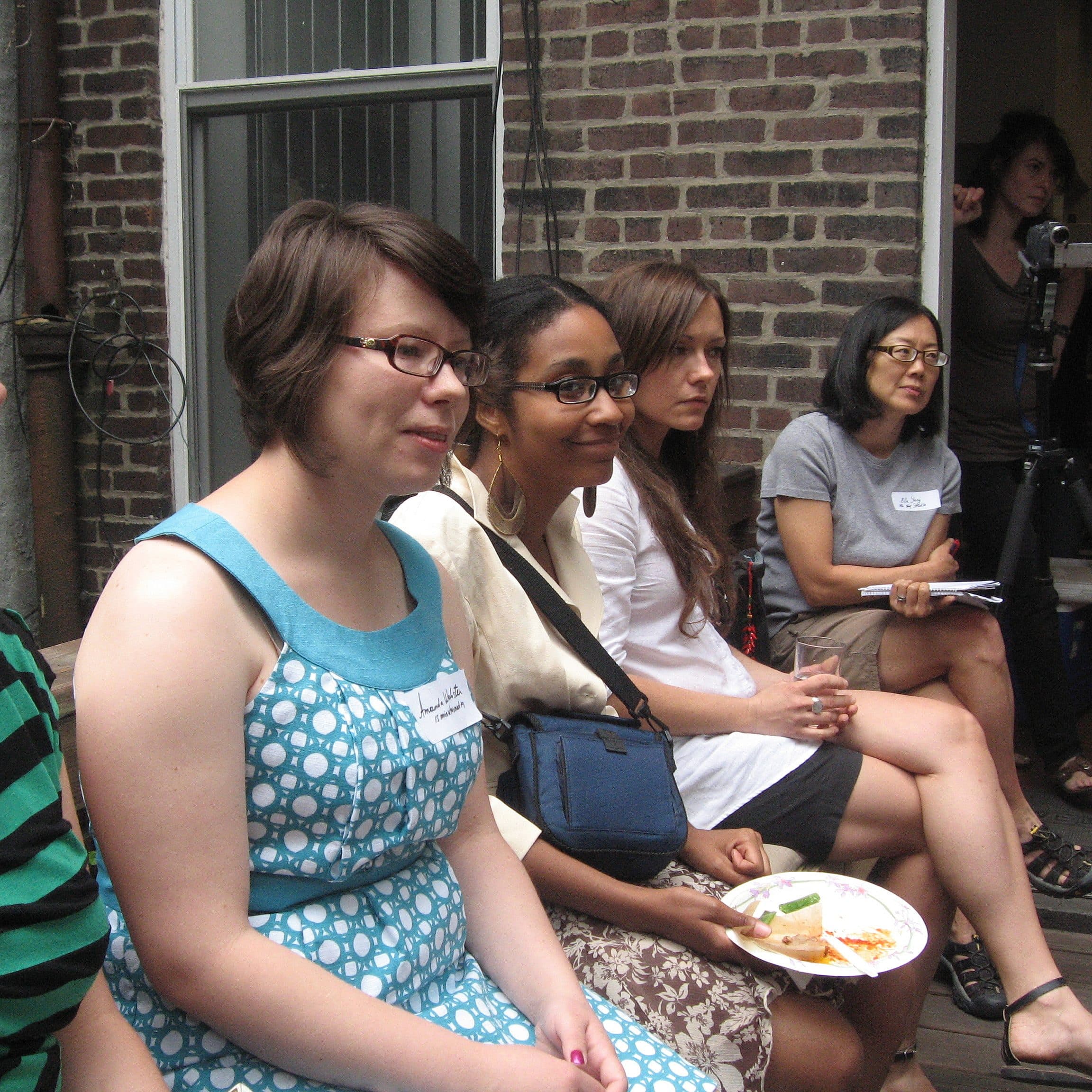 A group of people, primarily women, sitting closely together outdoors, some holding food plates. They are engaged in listening, with a brick wall and a person operating a camera in the background. The mood appears to be casual and attentive.