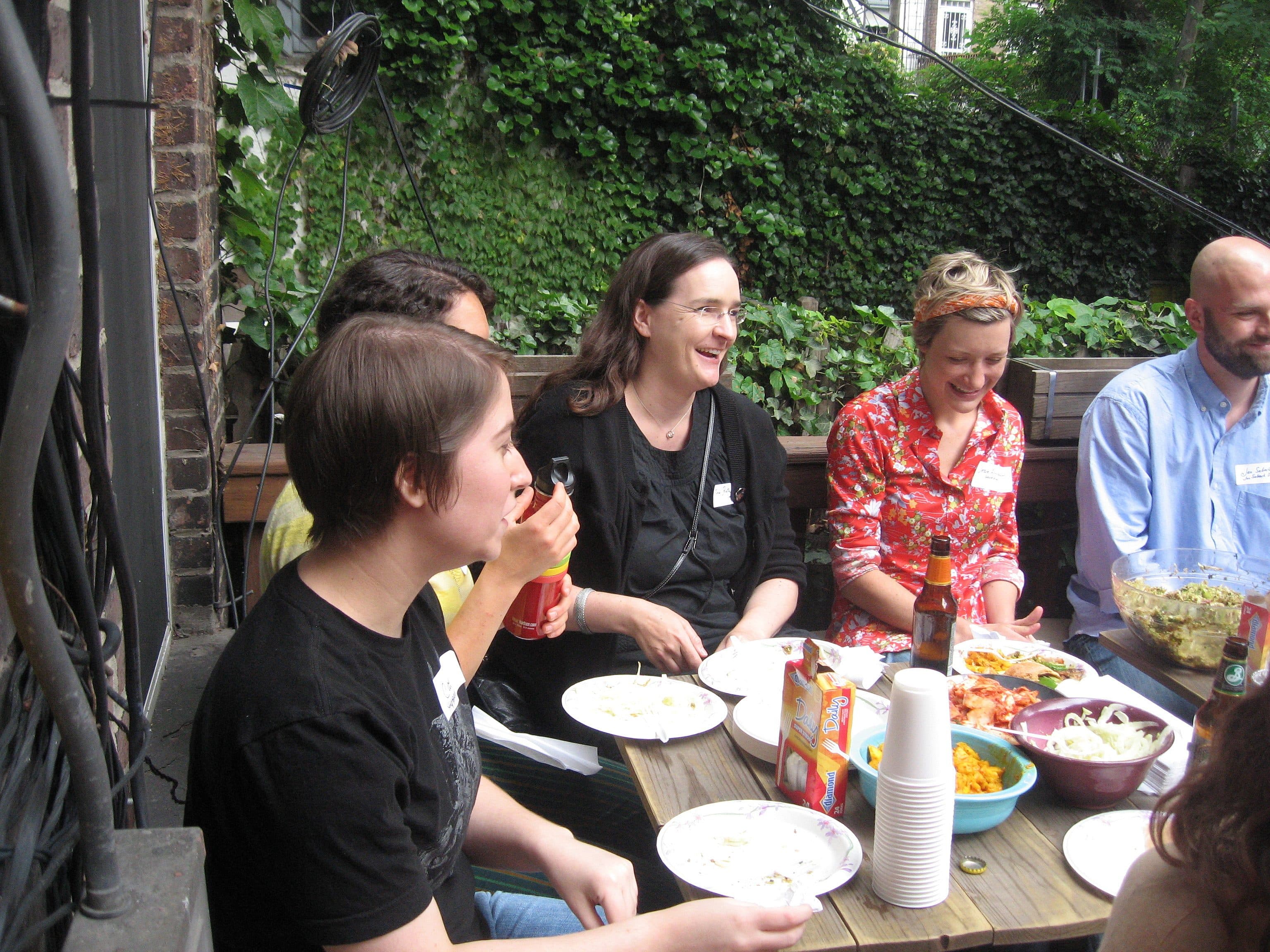 A group of people sitting around an outdoor table covered with plates of food and drinks. They are talking and smiling. There is a lush green wall of ivy in the background. The table has various dishes, cups, and bottles.