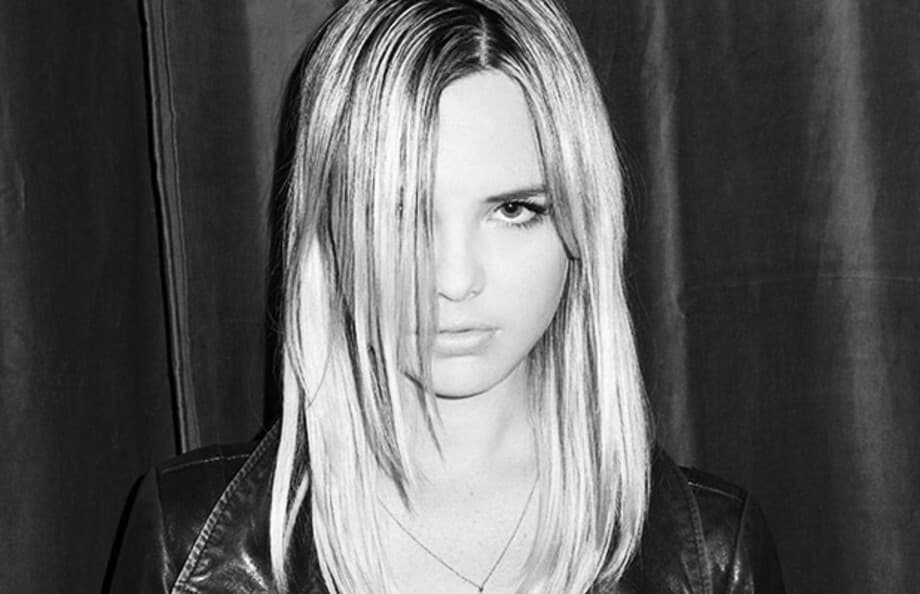 Black and white photograph of a person with long, straight, light-colored hair. One side of their hair partially covers their face. They are wearing a leather jacket and looking directly at the camera with a neutral expression. The background is a plain, dark curtain.