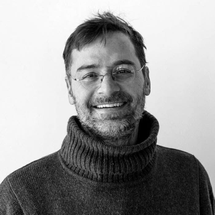 A smiling man with glasses and a beard is wearing a dark, thick, ribbed turtleneck sweater. He has short hair and stands against a plain white background. The image is in black and white.
