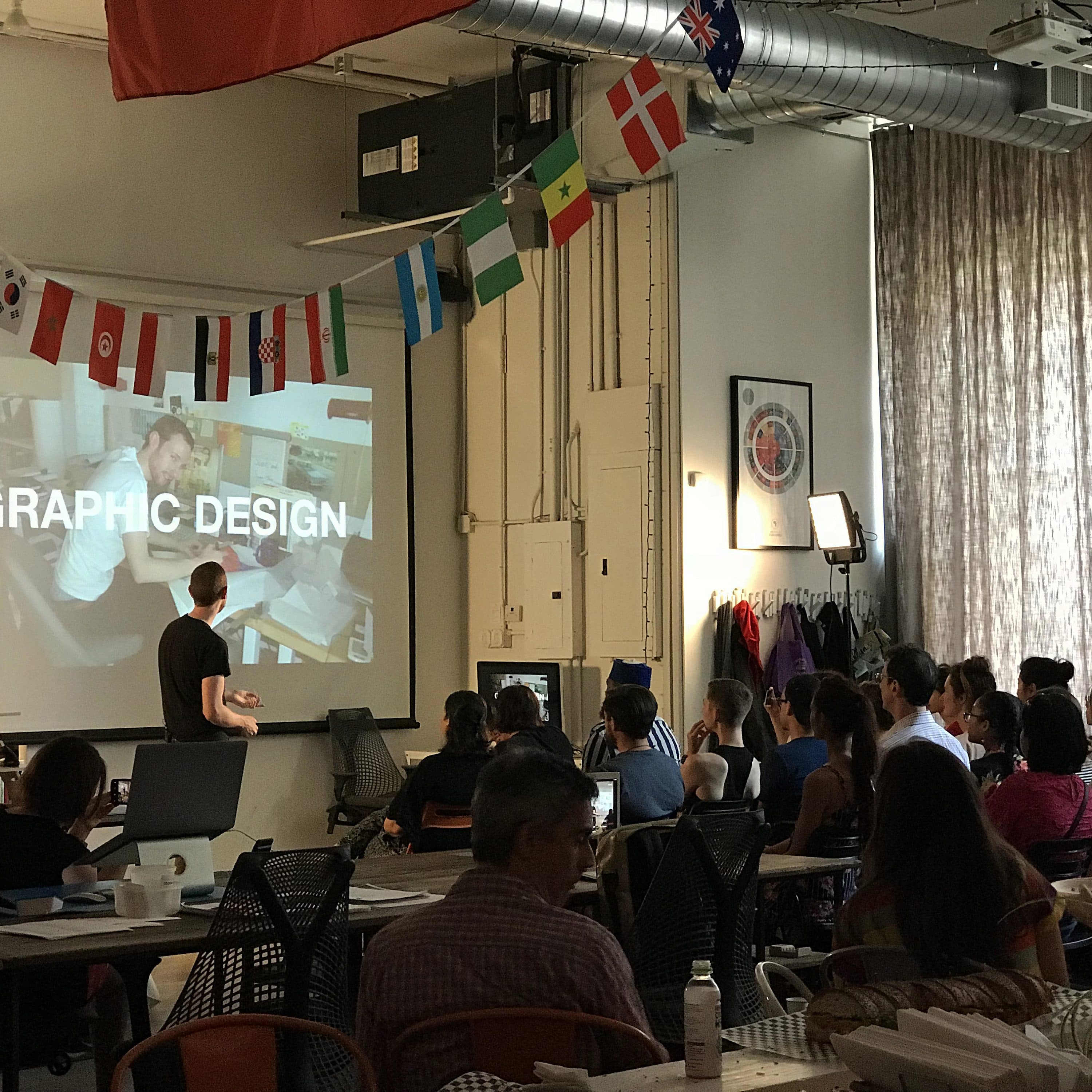 A person stands in front of a large screen giving a presentation about graphic design to a seated audience in a spacious room. The room is decorated with various international flags hanging from the ceiling, and long curtains cover the windows.