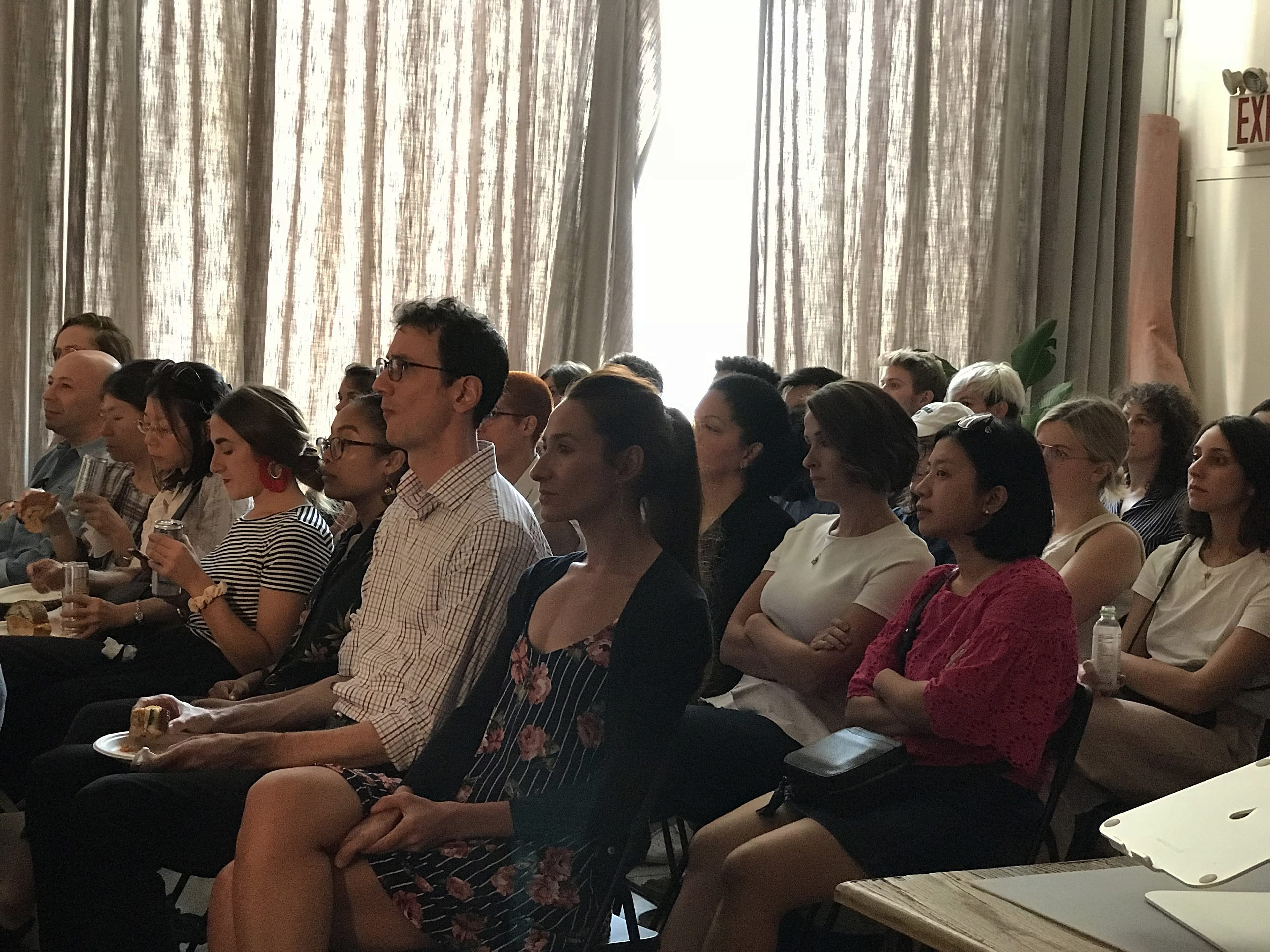 A diverse group of people sits closely together in a dimly lit room, attentively watching something off-camera. Some hold drinks or snacks, and several are taking notes. The room is illuminated by light filtering through curtains at the back.