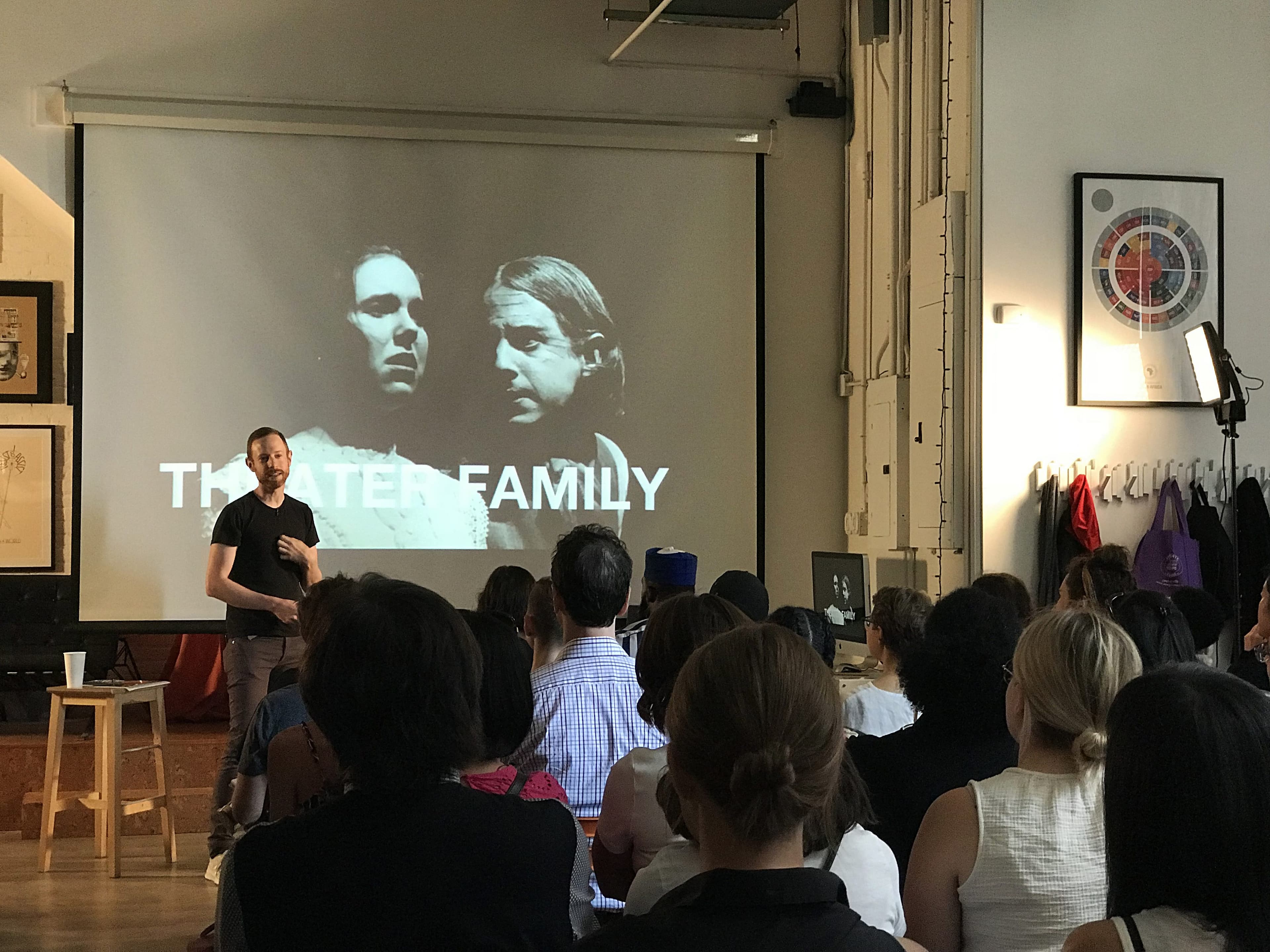 A man is giving a presentation to a seated audience in a well-lit room. Behind him is a large projector screen displaying a slide with black-and-white images of two people and the text "THEATER FAMILY." Several pieces of artwork are hung on the walls.