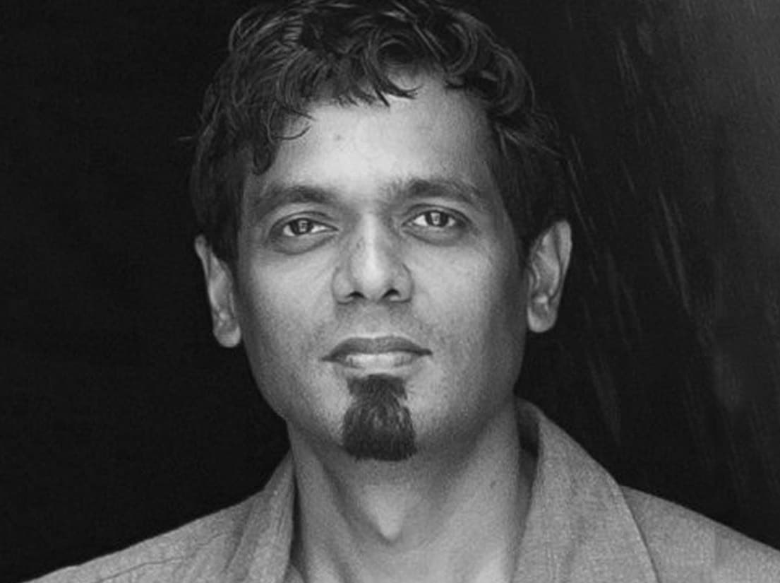 Black and white portrait of a person with short, wavy hair and a goatee. They are looking directly at the camera with a neutral expression. The background is dark, making the face stand out prominently.