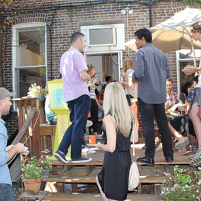 A group of people are gathered on a wooden deck outside a brick building, socializing. Some are chatting while others are holding drinks. Potted plants line the edges of the deck. An open door leads inside, and the area is lit naturally by daylight.