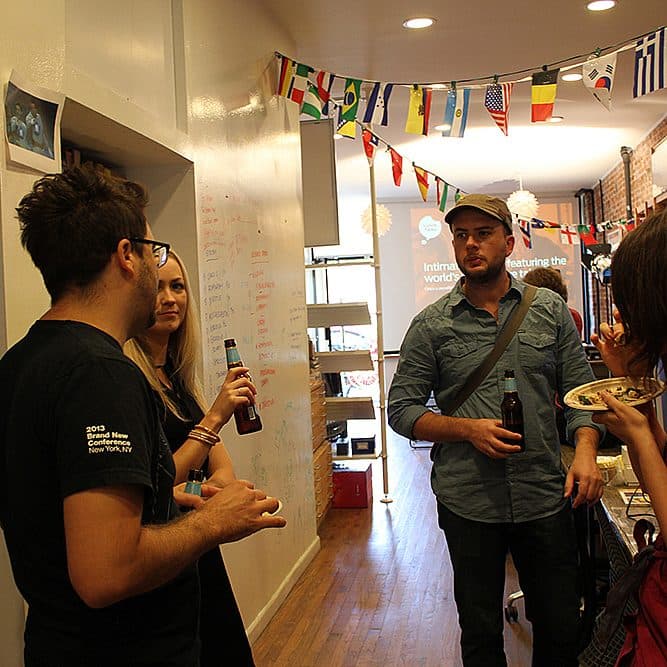 Four people are talking and drinking in a narrow, well-lit room decorated with international flags hanging from the ceiling. One person is eating from a plate, while the others hold drinks. The walls have various items, including notes written on a whiteboard.