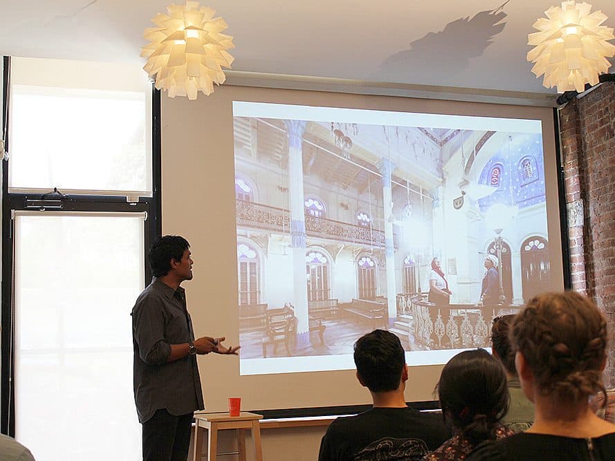 A person is giving a presentation to an audience in a bright room. The screen behind displays an image of an interior space with ornate architecture. The audience members are seated and focused on the speaker. Pendant lights hang from the ceiling, and exposed brick walls are visible.
