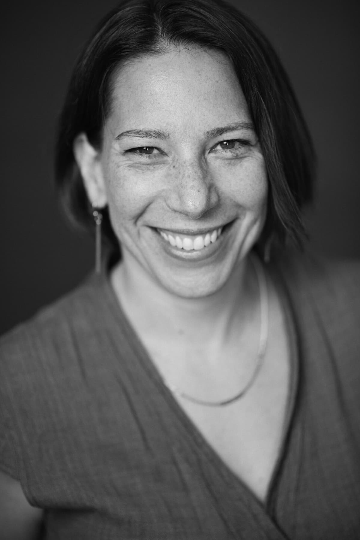 Black and white portrait of a smiling person with short, dark hair. They are wearing a V-neck top, a thin, curved necklace, and a single long earring. The background is plain and slightly blurred, emphasizing their bright and warm expression.