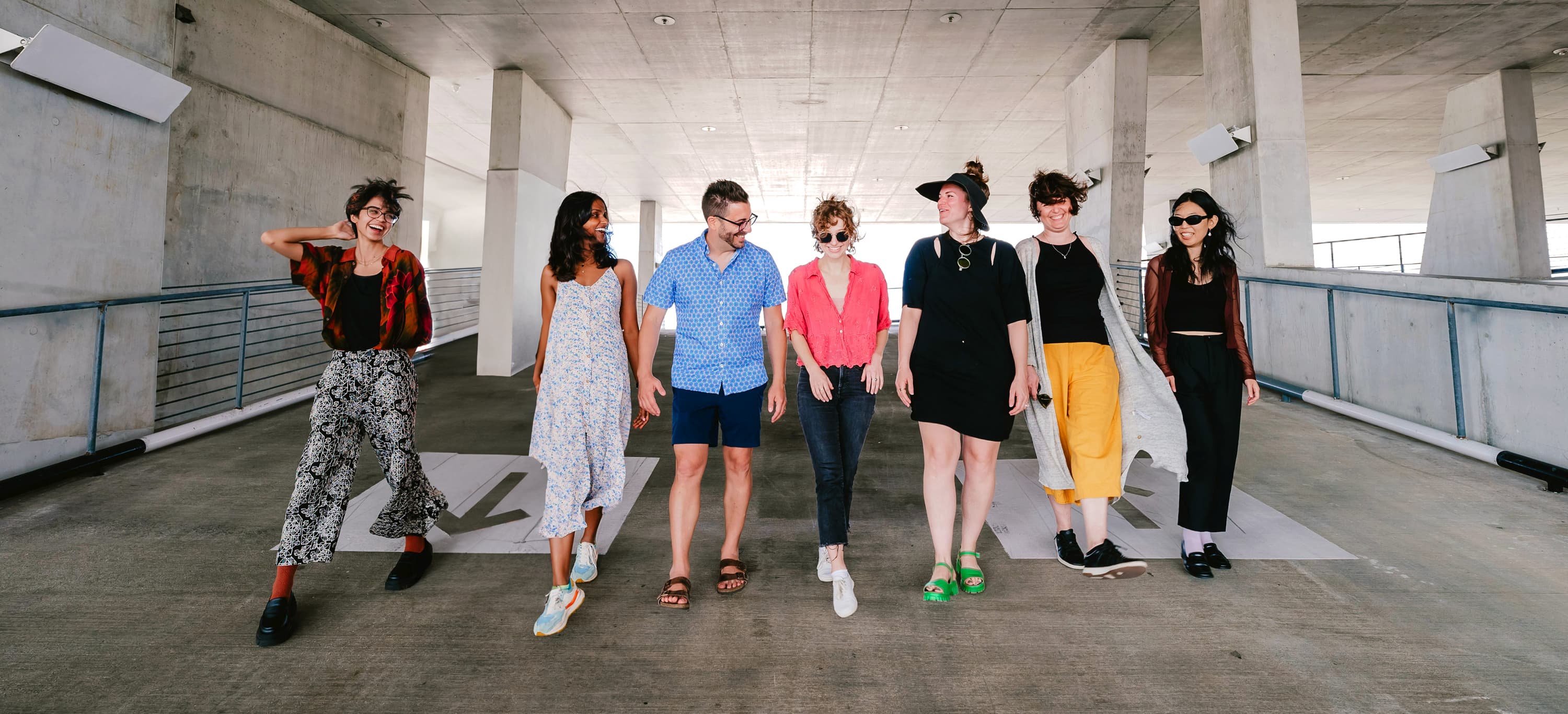 A diverse group of seven people walks together in a spacious, concrete structure, smiling and chatting. They are dressed in various casual and colorful outfits, ranging from shorts and t-shirts to dresses and jumpsuits. The setting appears to be an urban location.