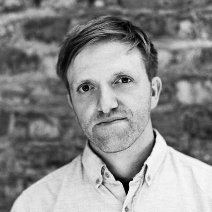 A person with short hair and light facial hair, wearing a button-up shirt, is looking directly at the camera. The background consists of a textured brick wall. The image is in black and white.