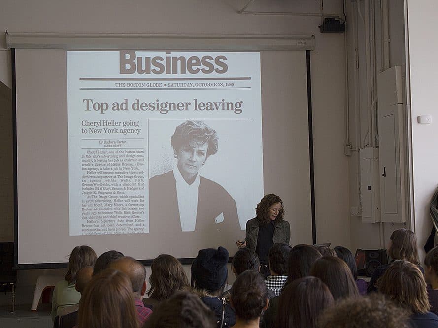 A speaker stands in front of an audience delivering a presentation. A newspaper clipping is projected on the screen behind them, with the headline "Top ad designer leaving." The room is filled with attendees seated and attentively listening.