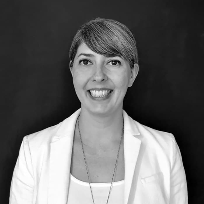 Black and white portrait of a person with short hair, smiling, wearing a light-colored blazer over a white top, and a long necklace. The background is plain and dark, emphasizing the person's friendly expression.