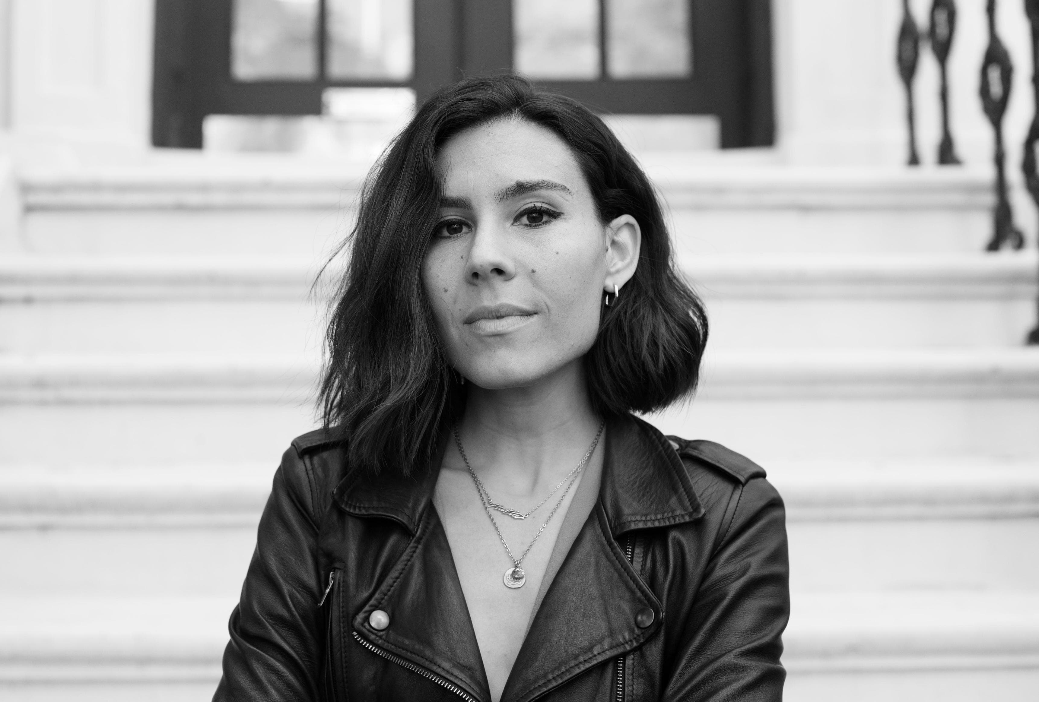 Black and white photo of a person with shoulder-length hair, wearing a leather jacket and layered necklaces, standing outside in front of white steps and black doors. They look confidently at the camera with a neutral expression.